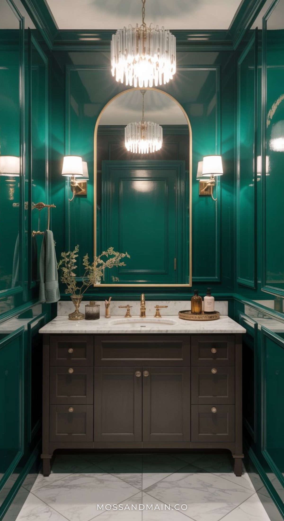Elegant luxe bathroom with glossy teal paneled walls, a gold-framed oval mirror, marble countertop, dark vanity, and luxe gold fixtures. Twin wall sconces and a modern crystal chandelier create moody bathroom vibes. Decor and hand towels adorn the counter.