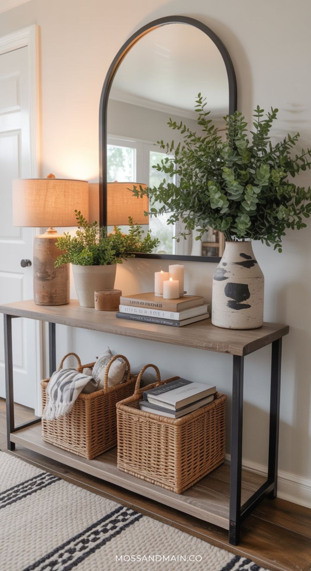 A wooden console table serves as stylish entryway table decor, with a mirror above, a lamp, potted plant, candles, books, and two woven baskets on the lower shelf, all set against a light-colored wall.