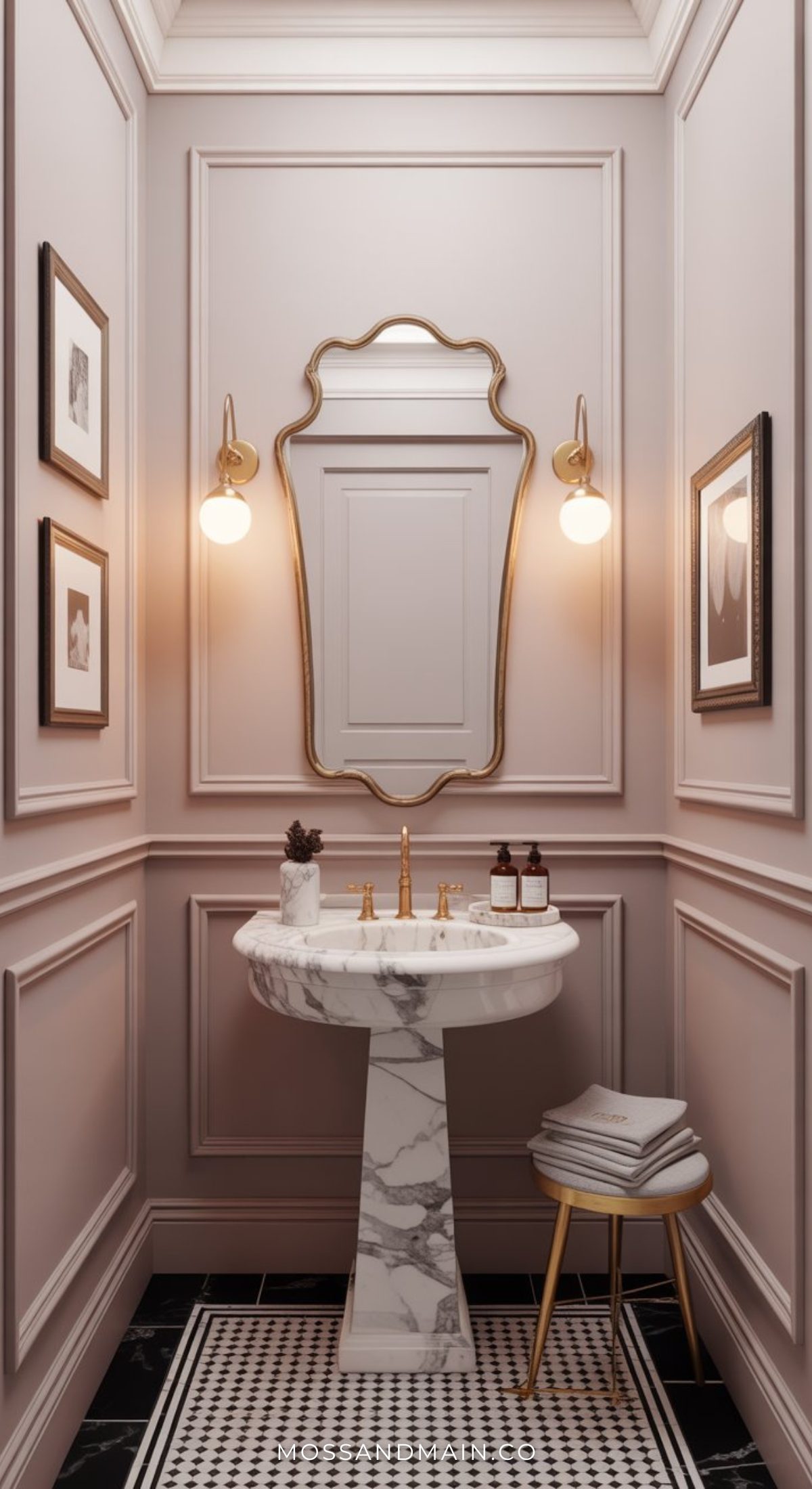 Elegant, narrow stylish bathroom with light pink paneled walls, a gold-framed mirror, two globe wall sconces, a marble pedestal sink, a gold faucet, framed art, black-and-white tiled floor, and a stool with folded towels.