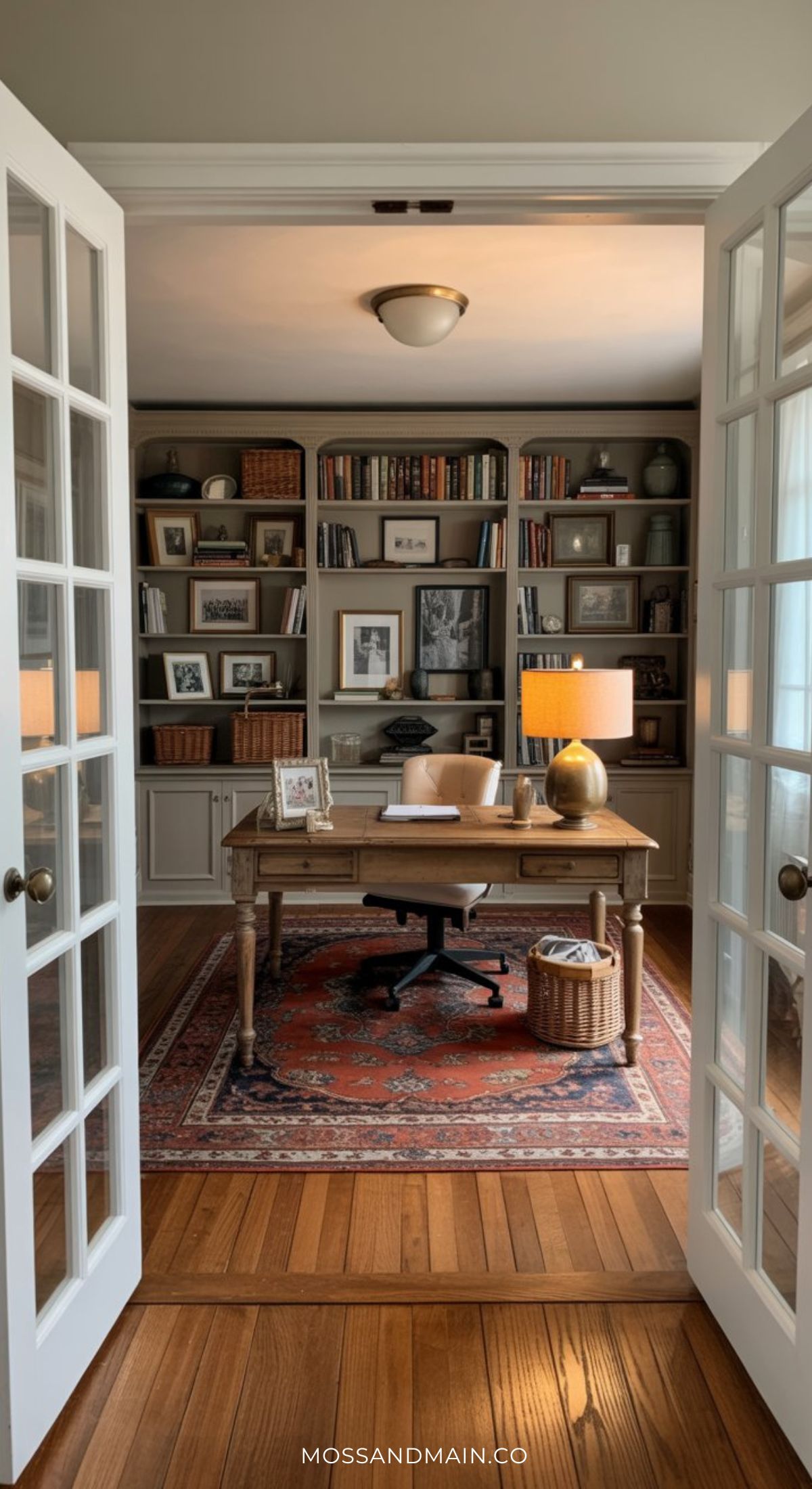 A cozy home office with a wooden desk, black chair, table lamps, and bookshelves filled with books and decor exudes a Nancy Meyers home aesthetic. French doors reveal the inviting space, while a patterned rug covers the wooden floor.