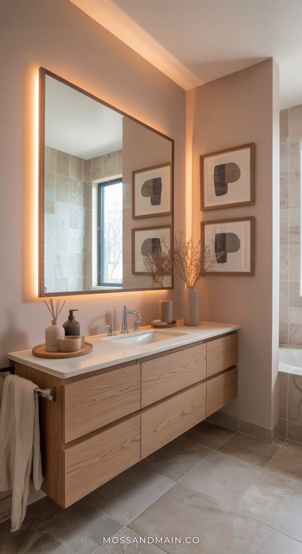 A stylish bathroom with a floating wood vanity, a large backlit mirror, minimalist decor, and beige tile walls. Two abstract framed artworks hang above the sink, adding bold bathroom flair to this modern space. Dried branches in a vase sit on the counter.