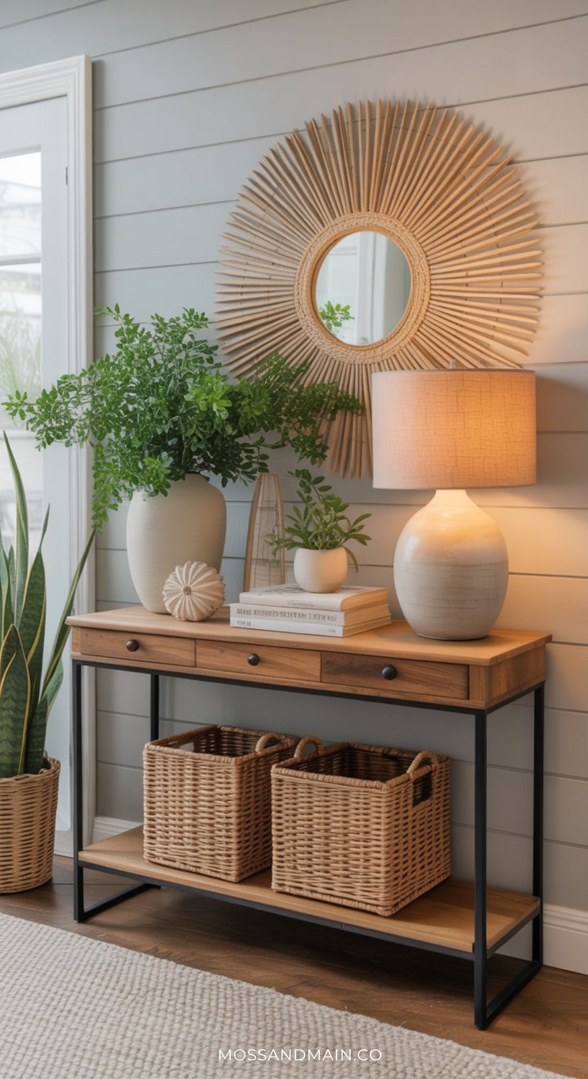 A modern entryway table decor scene with a wooden console, potted plants, stacked books, a ceramic lamp, two wicker baskets below, and a round woven mirror on a light gray paneled wall.