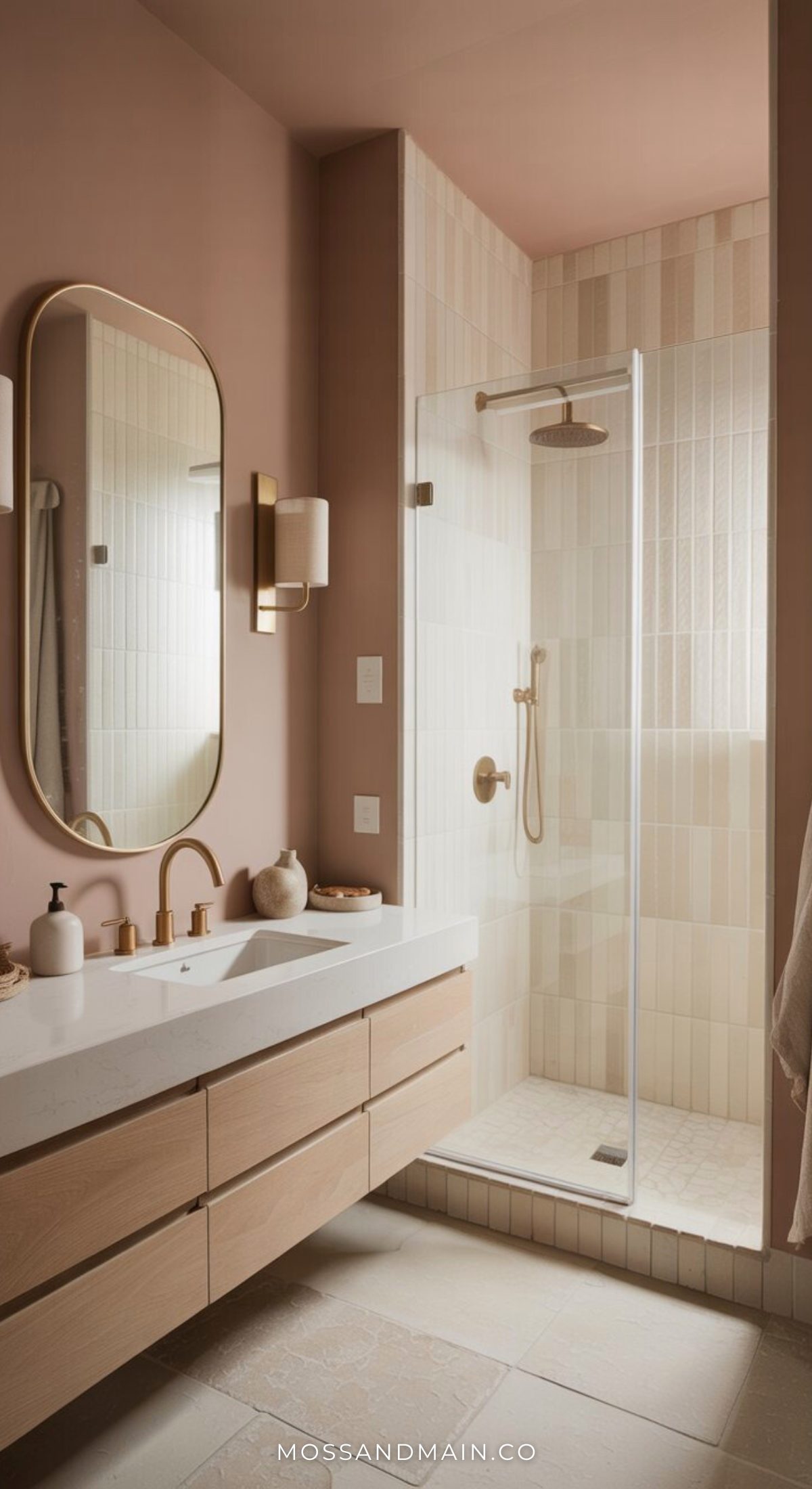 A modern bathroom with a light wood vanity, white countertop, oval mirror, and gold fixtures showcases bold bathrooms style. Glass-enclosed shower with beige tile and soft pink walls create a warm, inviting atmosphere—perfect for pink bathroom ideas.