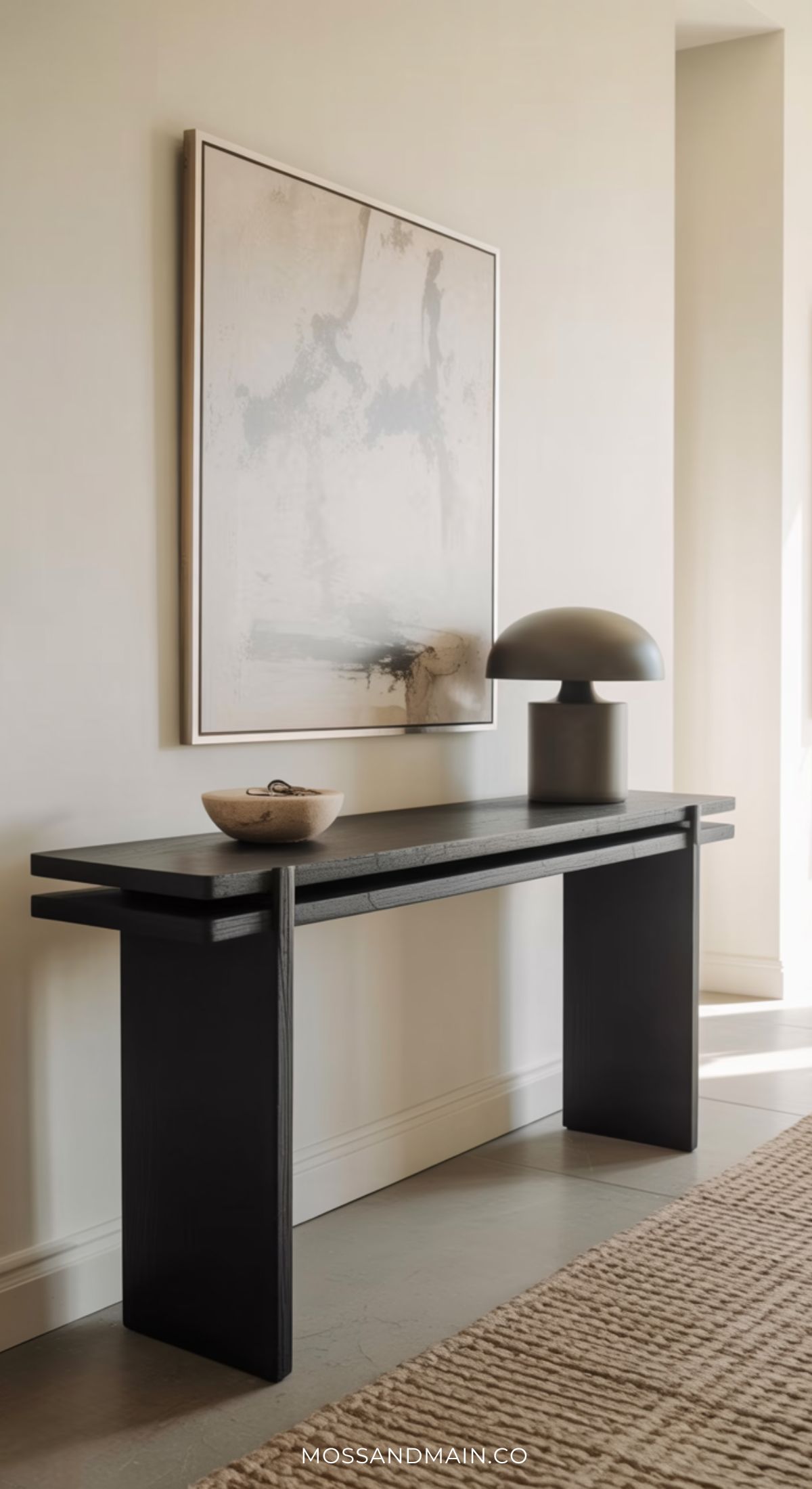A minimalist hallway features entryway table decor with a black console table, a beige bowl, and a modern gray lamp beneath an abstract painting. Natural light streams in, highlighting a textured rug on the floor.