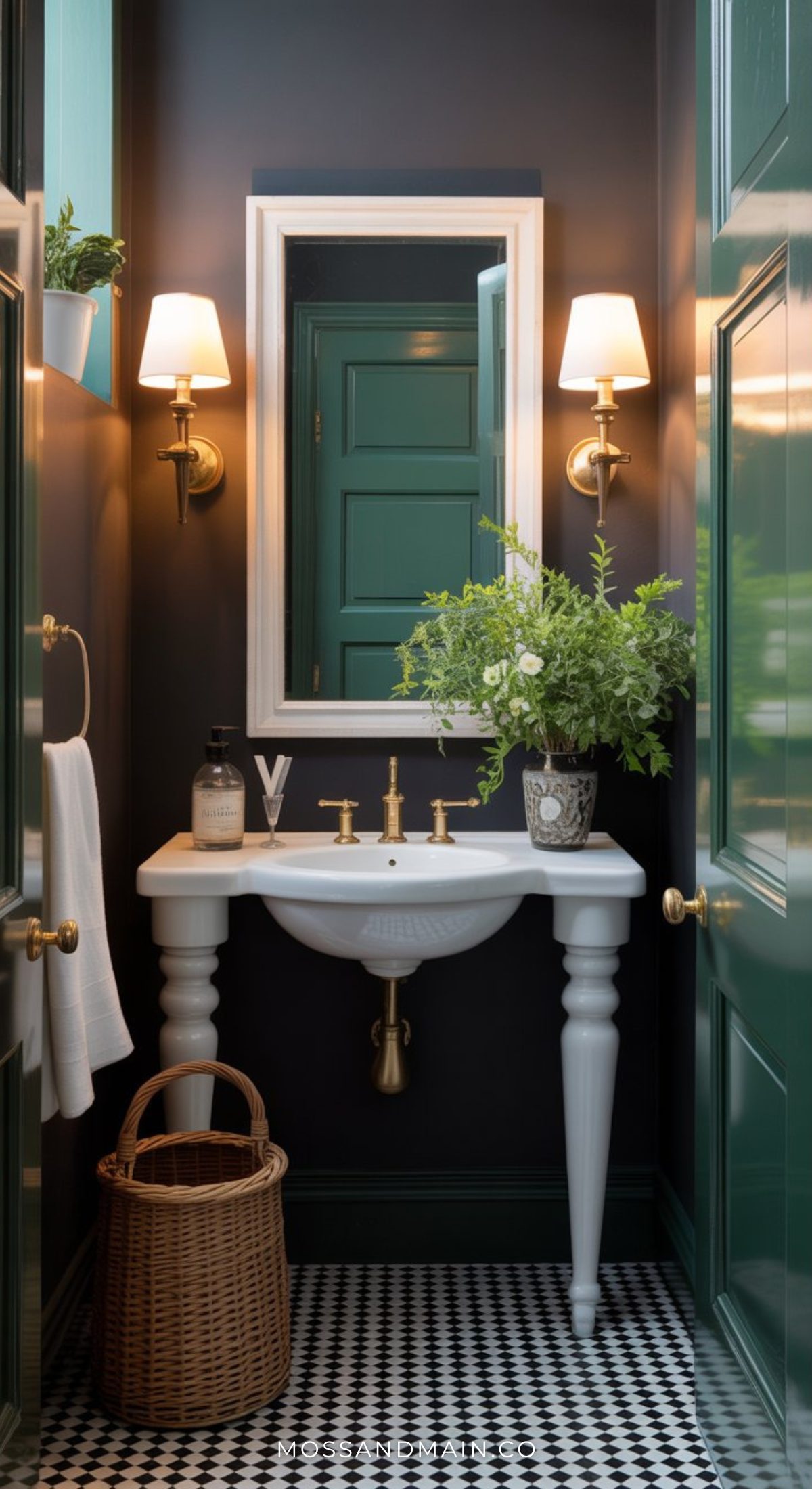 A small, elegant dark green bathroom with dark walls, a white pedestal sink, gold fixtures, a wicker basket, wall sconces, a framed mirror, and a vase of green foliage on the sink. Features a green door and black and white tile floor.