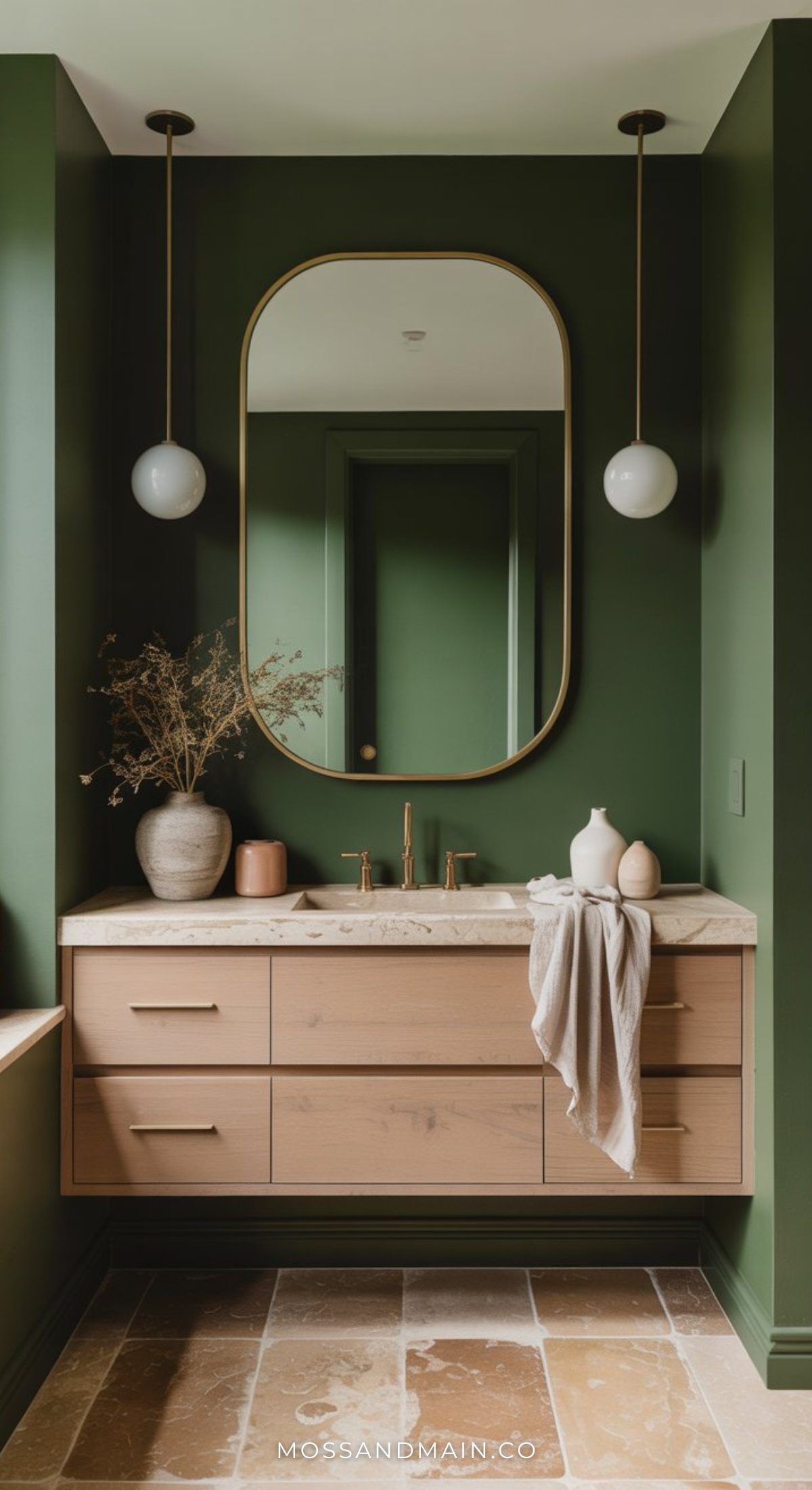 A moody bathroom with olive green walls, a large oval mirror, light wood floating vanity, marble countertop, ceramic vases, dried flowers, two globe pendant lights, and a brown tile floor.