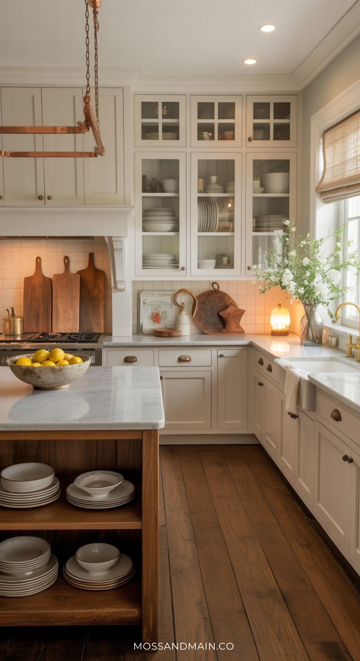 A bright, elegant kitchen with white cabinets, marble countertops, and glass-front cupboards radiates a Nancy Meyers home aesthetic—think wooden cutting boards, a bowl of lemons, and sunlight streaming through the window.