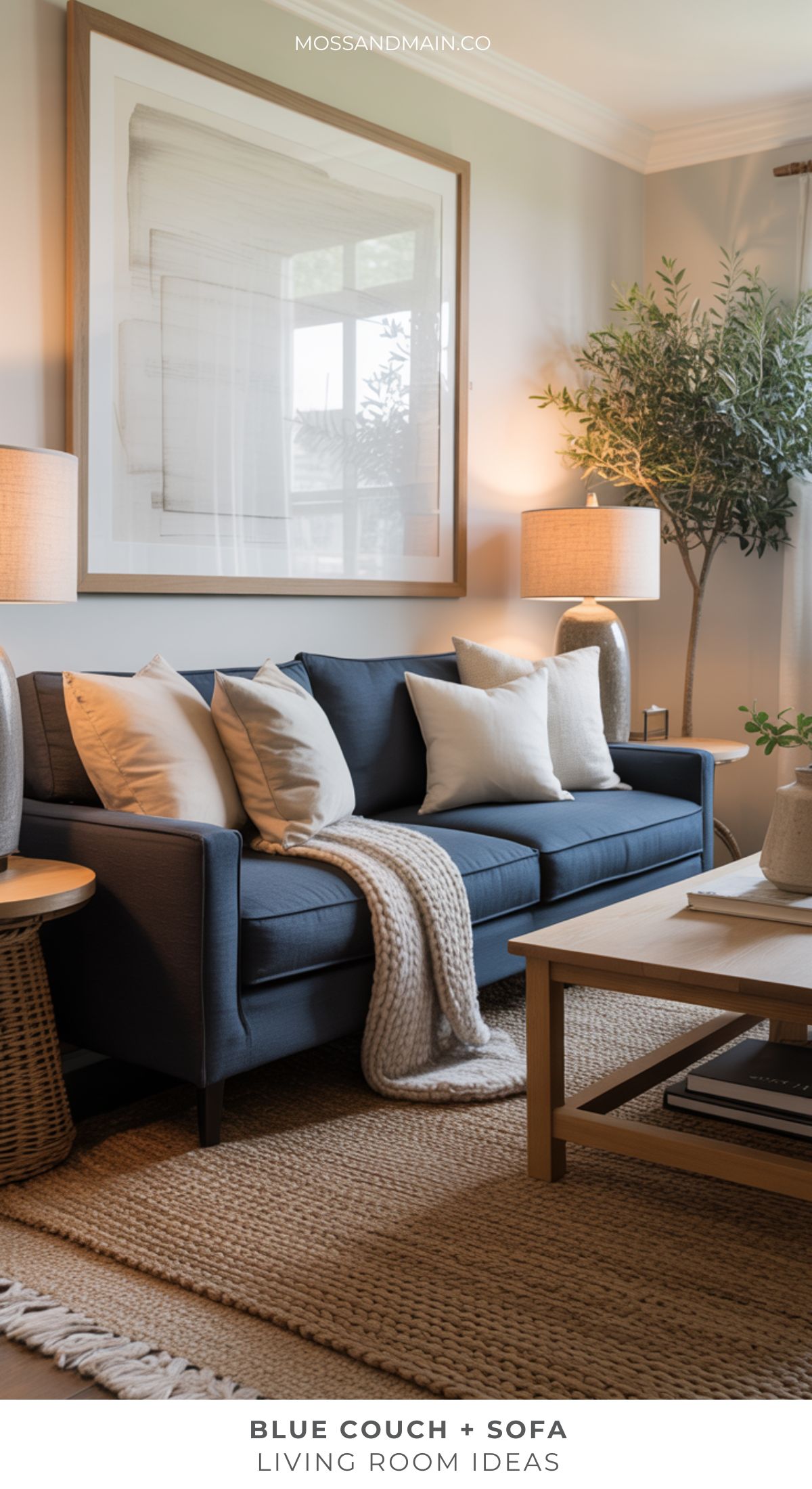 A cozy Blue couch living room featuring light throw pillows and a knit blanket, a wooden coffee table, woven rug, large framed wall art, side table with lamp, and a potted tree in the corner—perfect for fresh living room ideas.