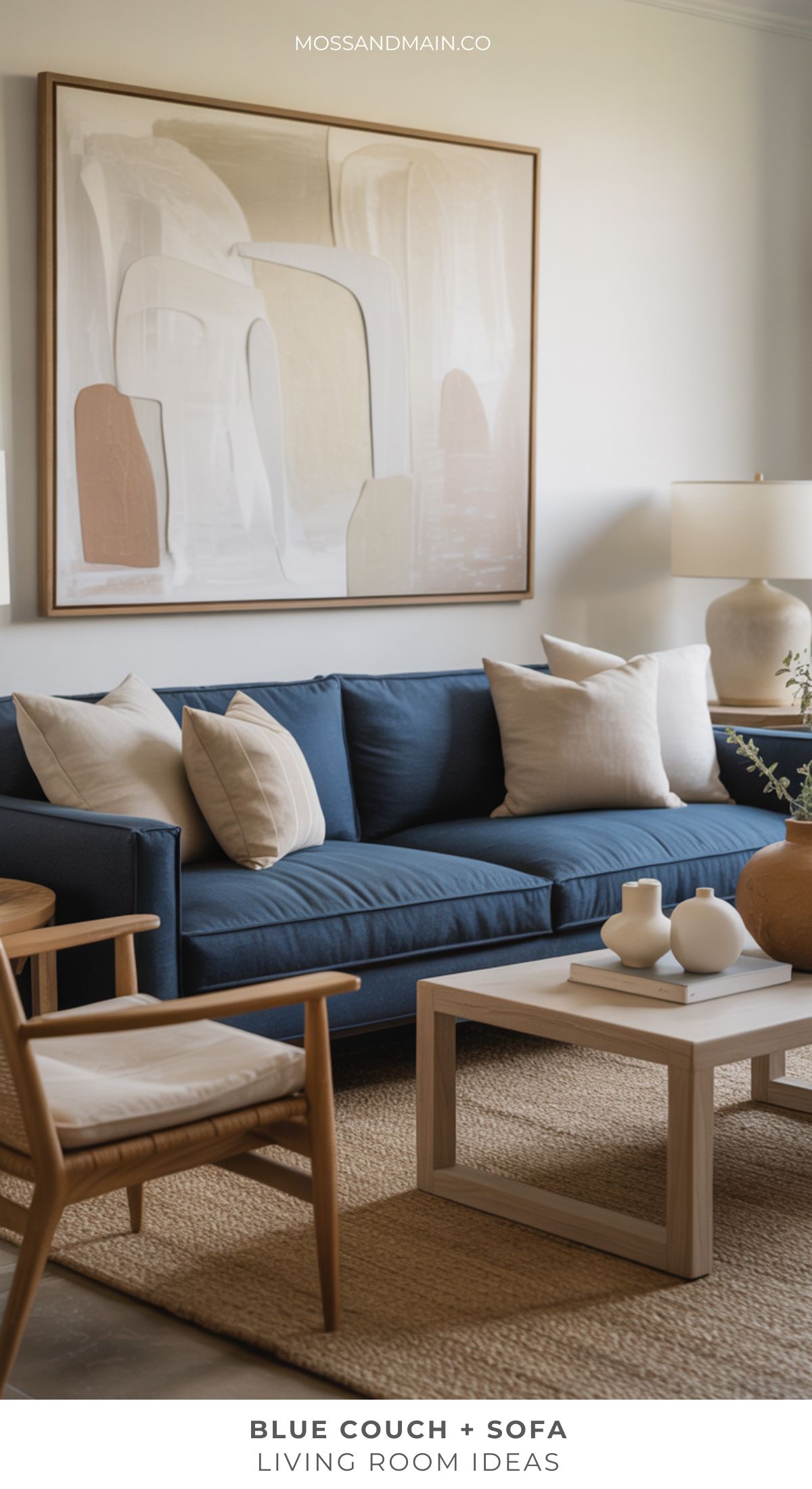 A modern blue couch living room featuring neutral pillows, a wooden armchair, white coffee table, textured rug, and abstract wall art above the sofa. A table lamp and decorative items complete the cozy space.