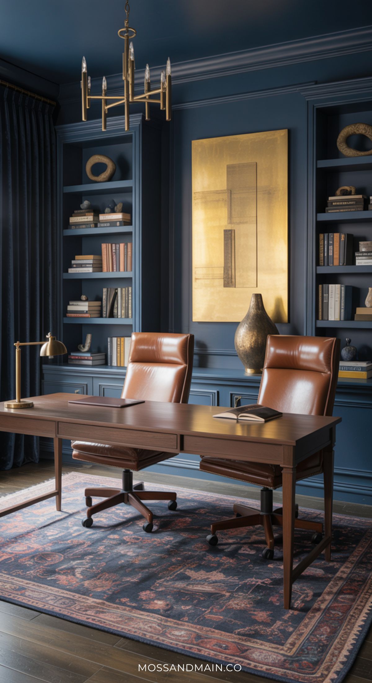 Elegant, moody home office with two brown leather chairs at a wooden desk, built-in bookshelves, a large gold abstract artwork, a decorative vase, and chandelier. Dark blue walls and a patterned rug complete this perfect home office for two.