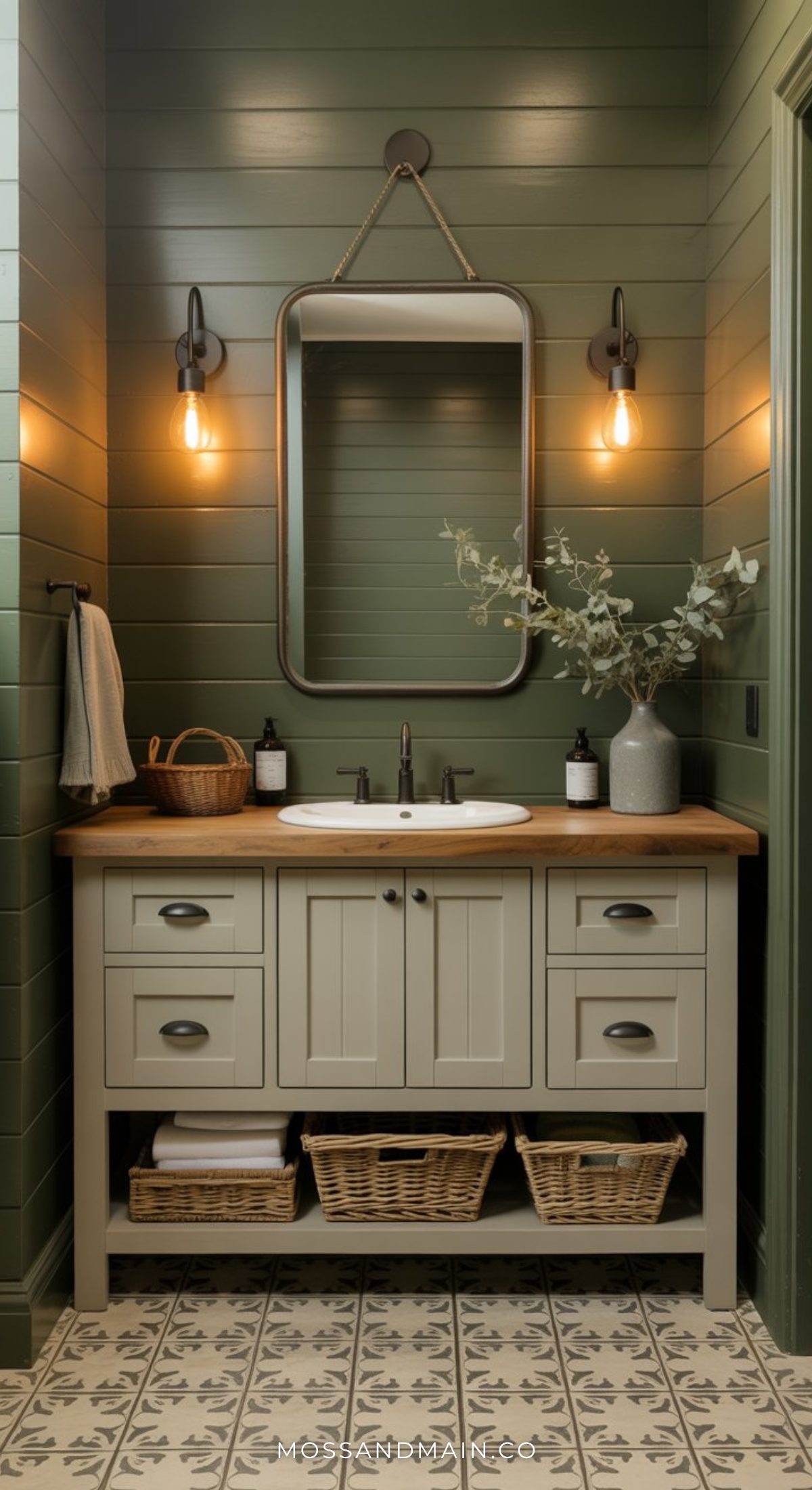 A moody bathroom with cozy green shiplap walls, a farmhouse-style vanity with wood countertop, wicker baskets, a rectangular mirror, and two wall sconces with exposed bulbs. Decorative tiles cover the floor.