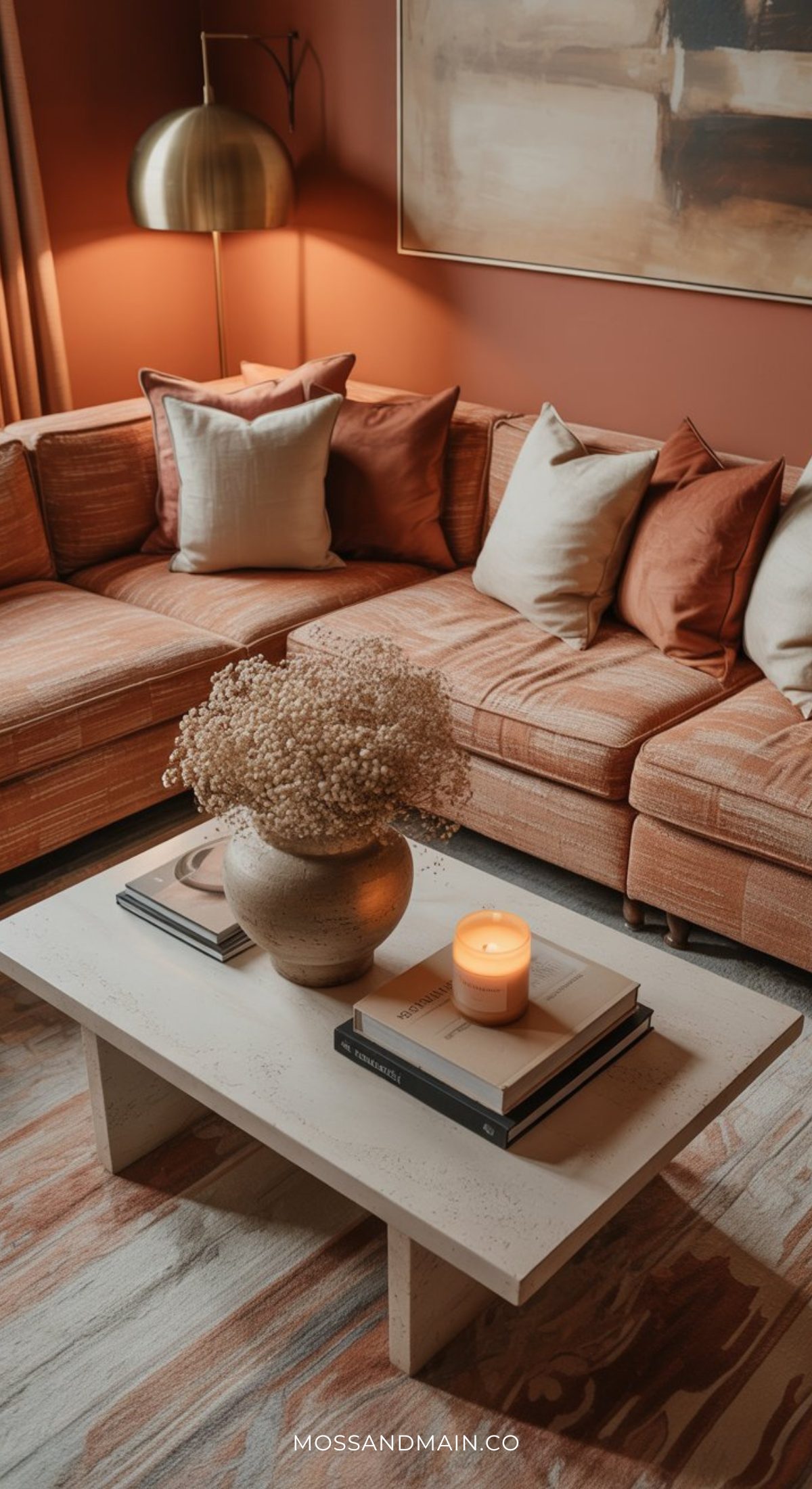 A cozy Soulful Home living room with a peach-colored sectional sofa, assorted throw pillows, a white coffee table holding dried flowers and a lit candle, books, a gold floor lamp, and warm-toned walls inspired by the Psychology of Color for 2026.
