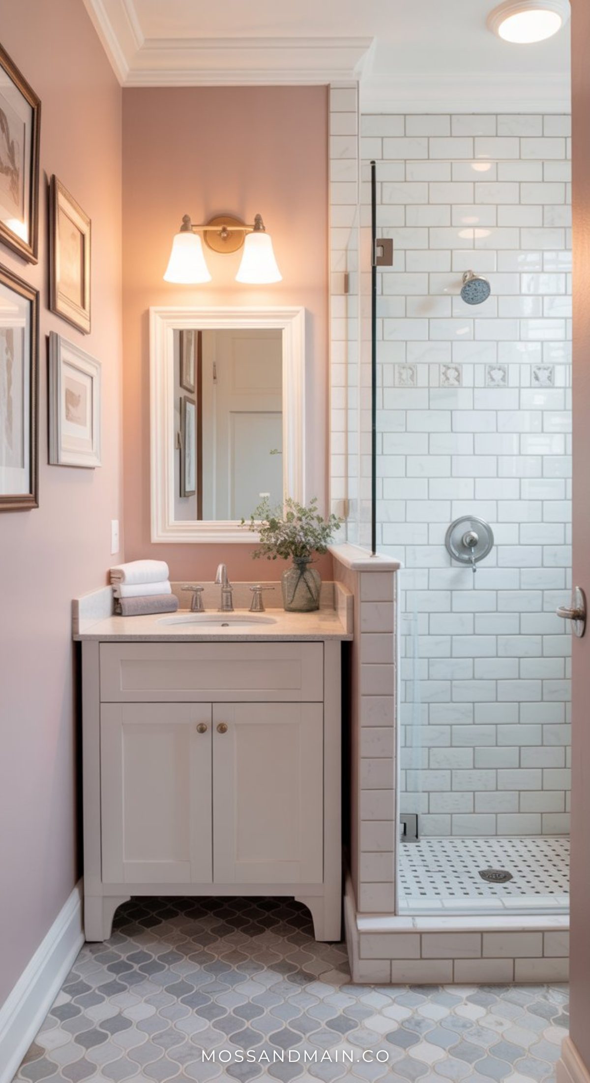 A small bathroom with mauve walls, a white vanity with sink and mirror, wall-mounted light, glass shower with white subway tiles, and patterned tile floor—perfect for those seeking pink bathroom ideas and bold bathroom decor. Flowers and rolled towels add charm.