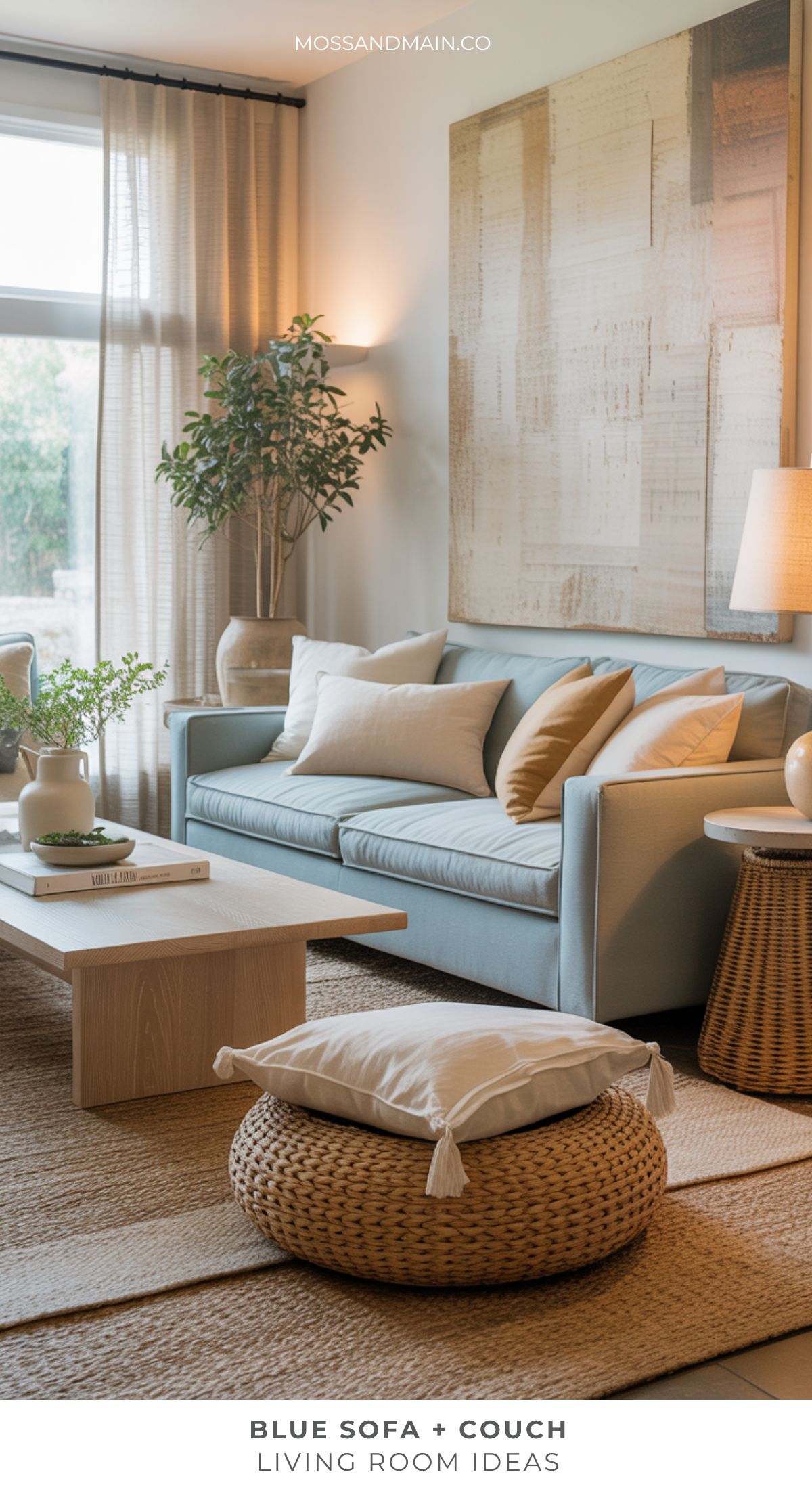 A modern blue couch living room featuring neutral throw pillows, a wooden coffee table, and a round woven pouf. Large art adorns the wall, while a plant and natural light stream in through sheer curtains.