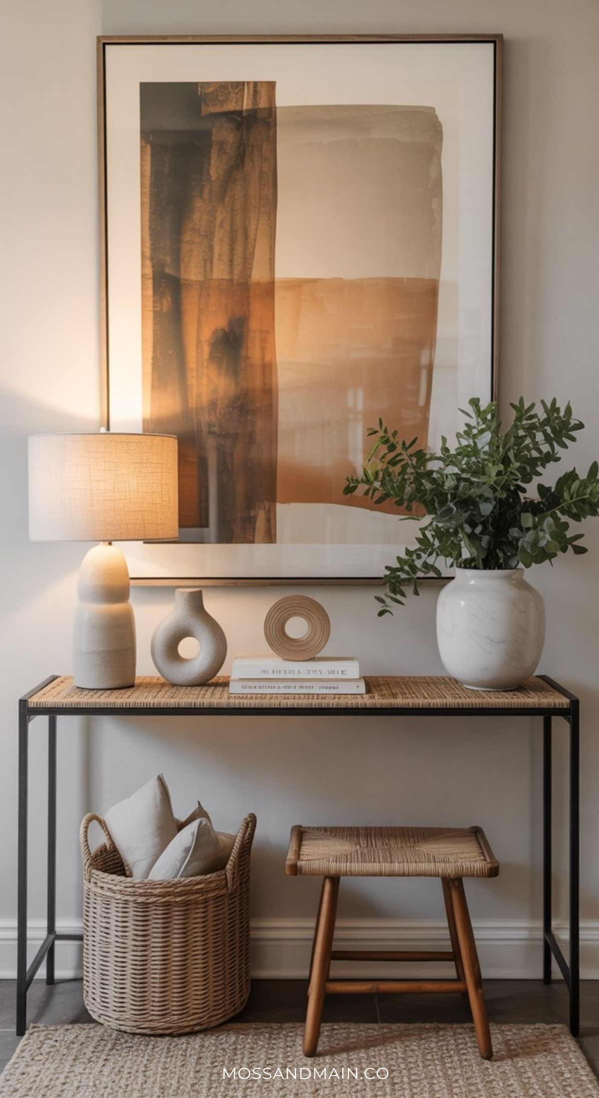 A modern entryway features stylish entryway table decor, including a textured lamp, ceramic accents, a vase with greenery, and framed abstract art. A small wooden bench and woven basket with pillows complement the look on a gray tile floor.