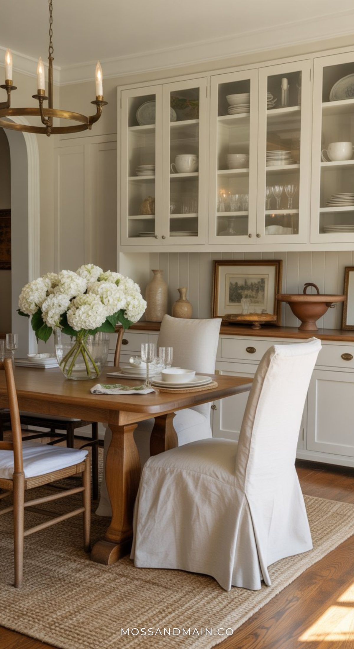 A cozy Nancy Meyers dining room with a wooden table set for four, white upholstered chairs, a vase of white flowers, and glass-door cabinets displaying dishes. Warm lighting and classic decor create an inviting, timeless atmosphere.