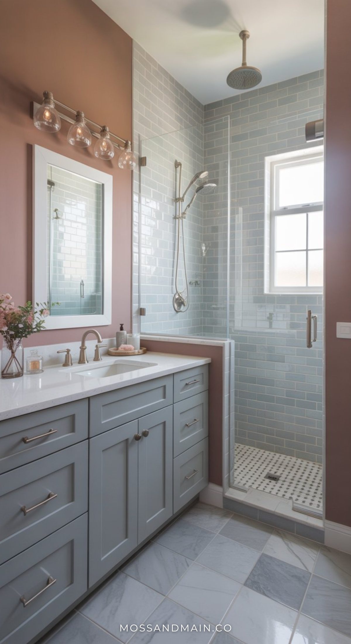 A stylish bathroom featuring a light blue vanity, white countertop, and glass-enclosed shower with gray tile. Above the sink, a mirror is framed by lights. Natural light flows in through the window onto the white and gray tile floor.