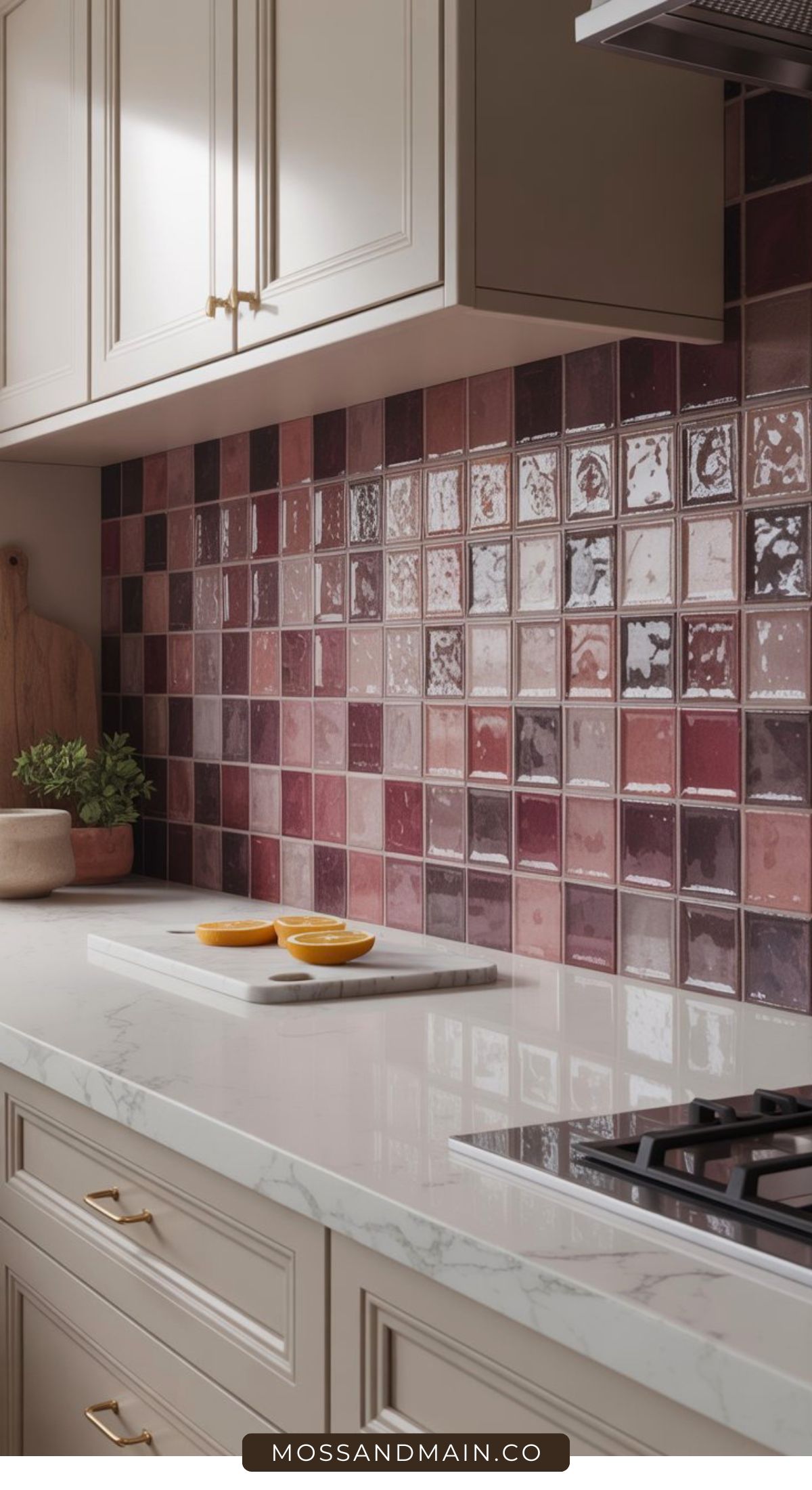 A modern kitchen with a marble countertop, beige cabinets, gold handles, and a gas cooktop. Glossy square tiles in burgundy and plum shades create a striking backsplash—a great space for exploring aubergine kitchen ideas. Two orange halves rest on a white cutting board.