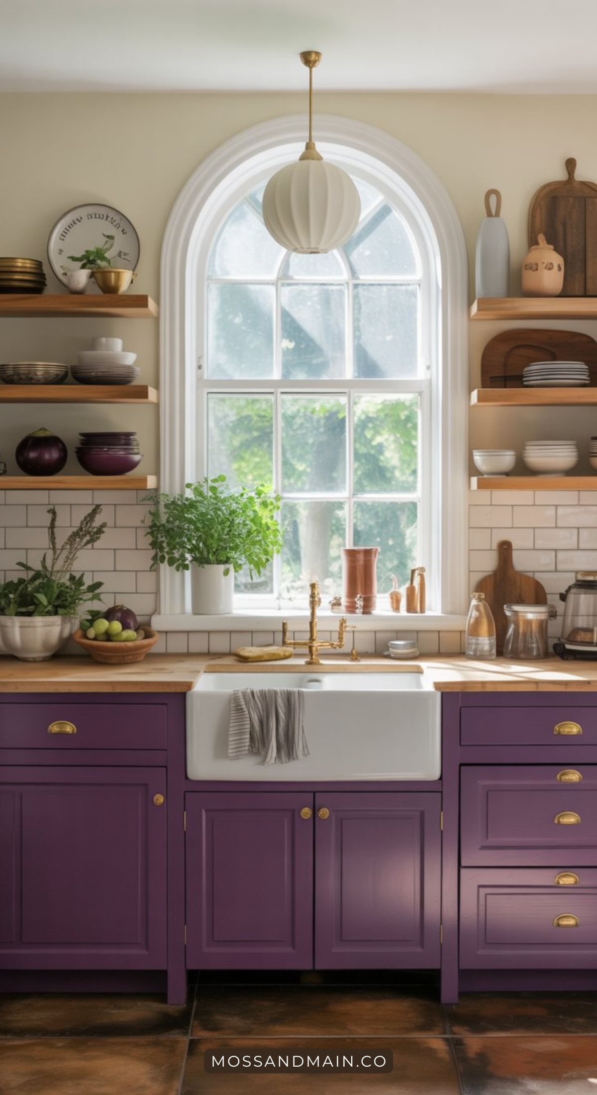 Bright kitchen design with purple cabinets inspired by Aubergine kitchen ideas, a white farmhouse sink, brass fixtures, wooden shelves with dishes and plants, and a large arched window. A towel hangs by the sink.