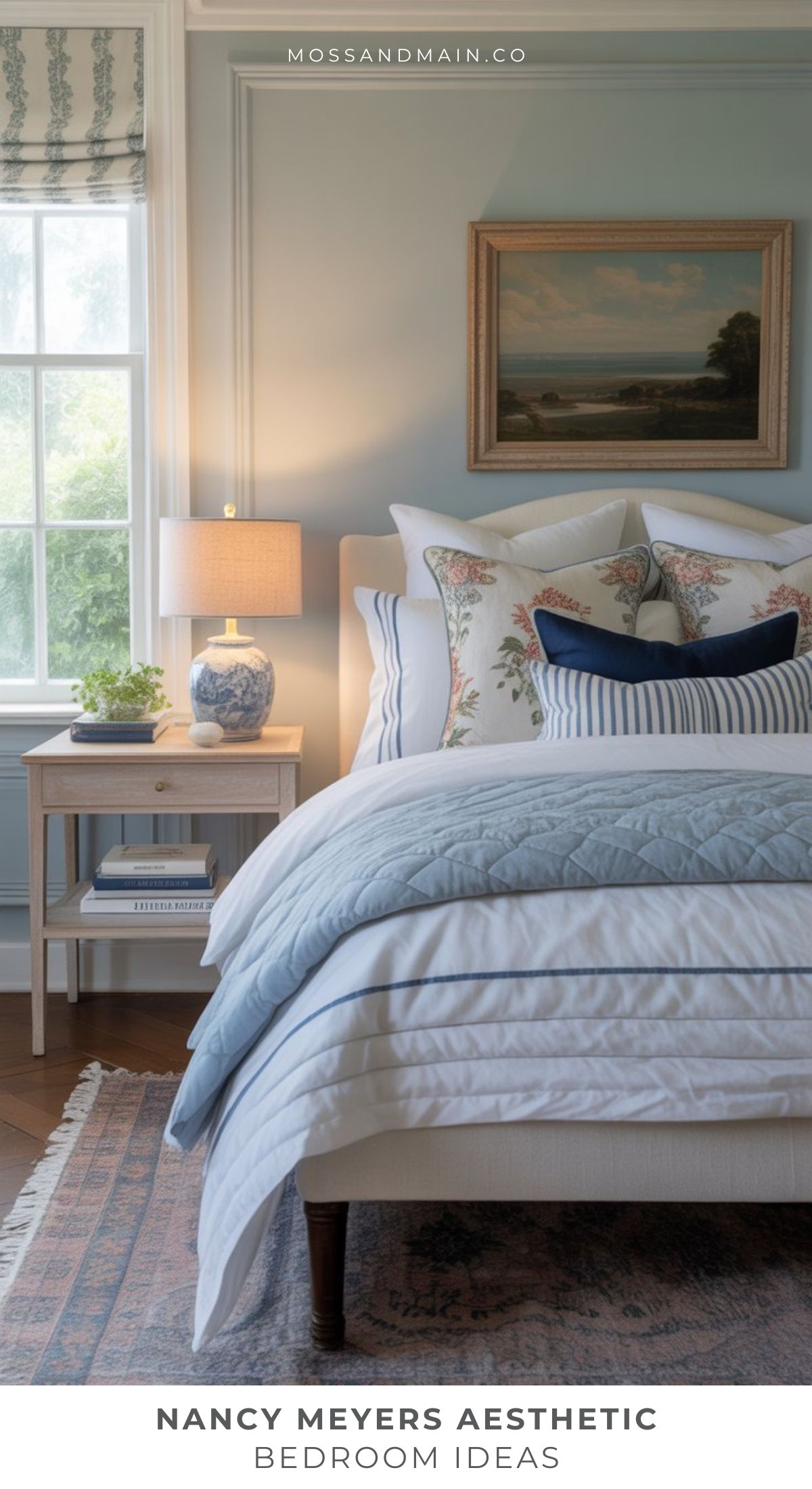 A cozy bedroom inspired by Nancy Meyers bedroom ideas, featuring light blue walls, a large window, plush white and blue bedding with floral pillows, a bedside table with books and lamp, a framed painting, and a classic patterned rug.
