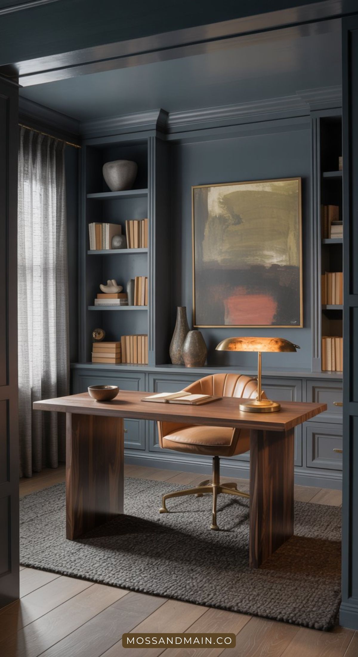 A modern home office with blue-gray walls features color drenching, built-in shelves of books and decor, a wooden desk, a leather chair, a brass desk lamp, and a large abstract painting above the desk. Light filters in through curtained windows.