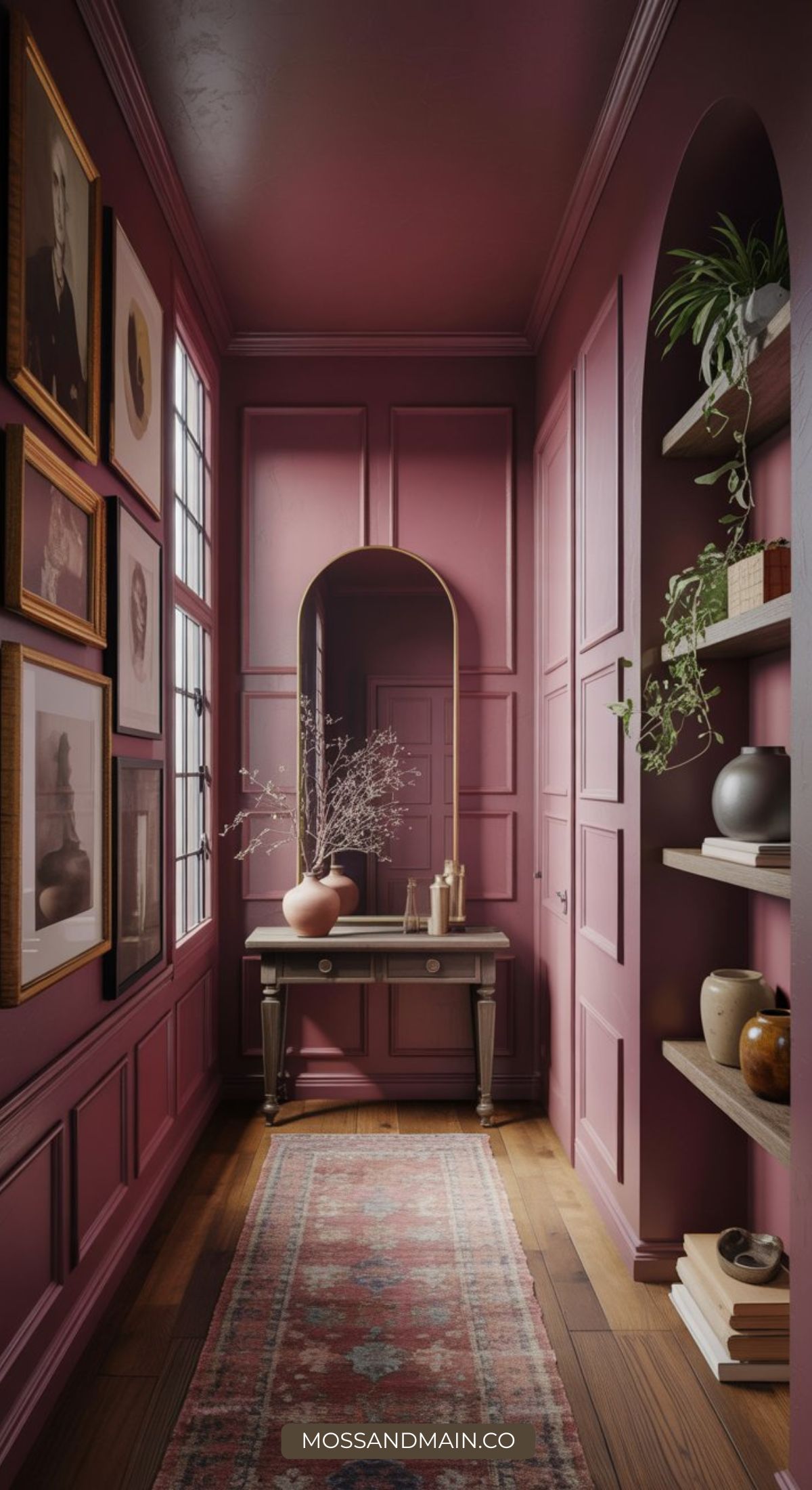 A narrow, color drenched hallway with mauve walls, wooden floor, and a patterned rug features framed art, a large arched mirror, a console table with vases, and built-in shelves with decorative objects and plants.