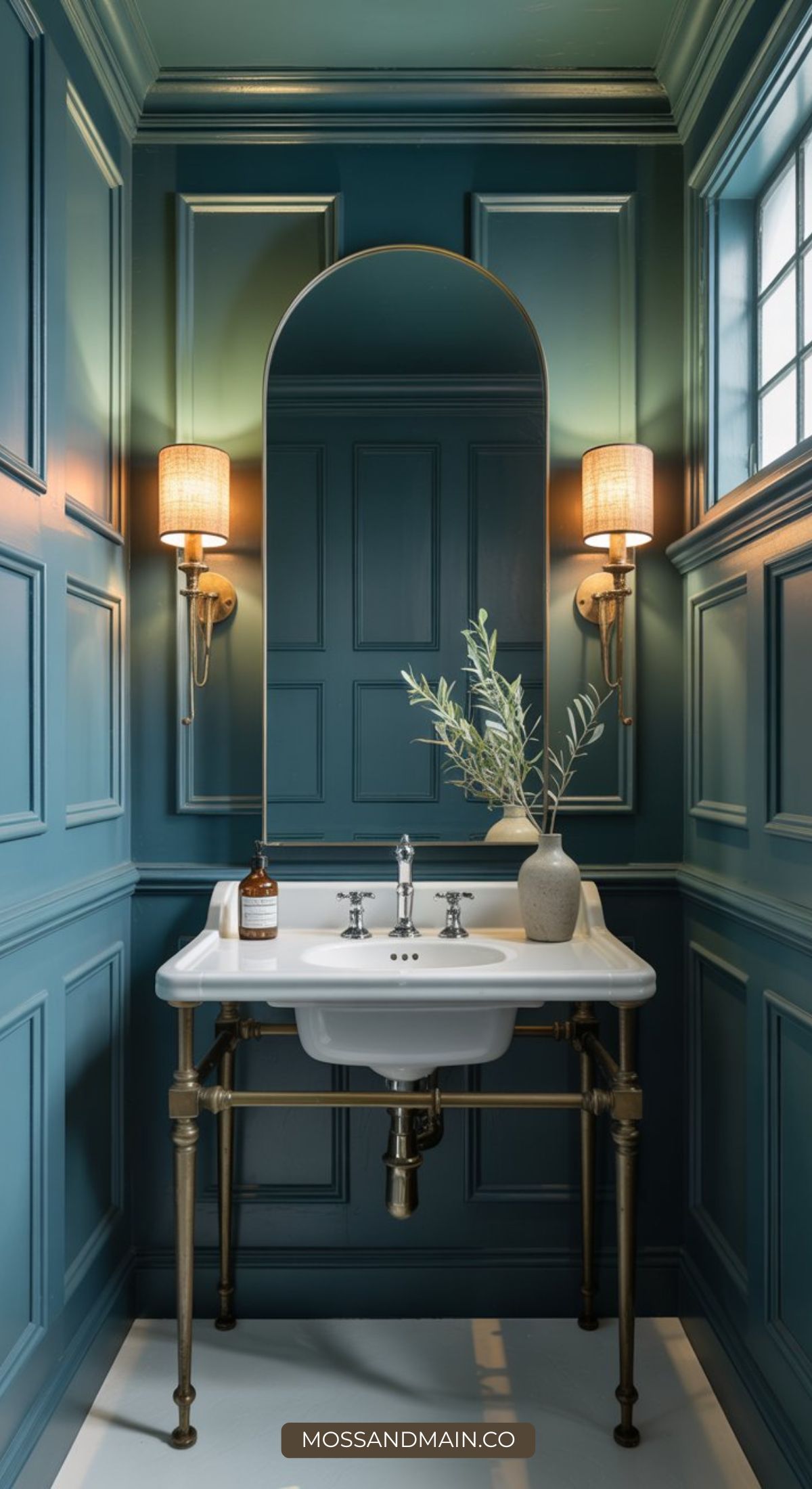 A stylish, color drenched bathroom with teal paneled walls, a white sink with brass legs, two wall sconces, a tall arched mirror, and decorative items like a soap bottle and a vase with greenery on the sink.