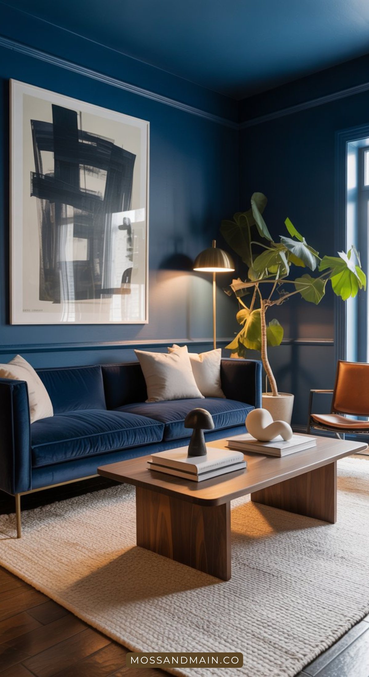 A modern, moody living room with dark blue walls, a velvet blue sofa, wooden coffee table with decor, large abstract art piece, tall floor lamp, potted plant, and a tan leather chair on a light textured rug—perfect for cozy living rooms.