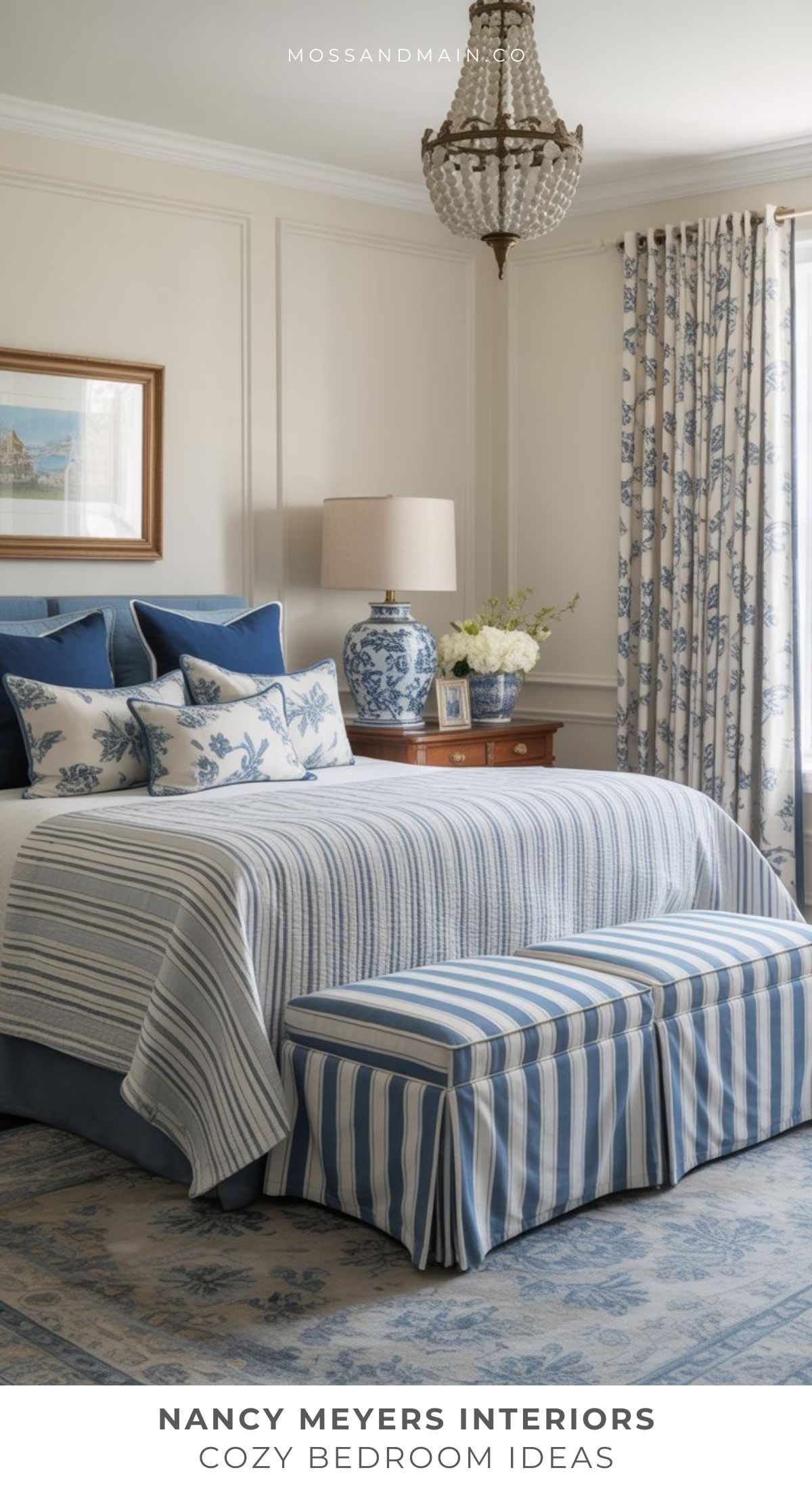 A cozy nancy meyers bedroom features a blue and white theme with striped bedding, floral pillows, a matching upholstered bench, a blue ceramic lamp, floral curtains, and a chandelier for that classic, elegant nancy meyers aesthetic.