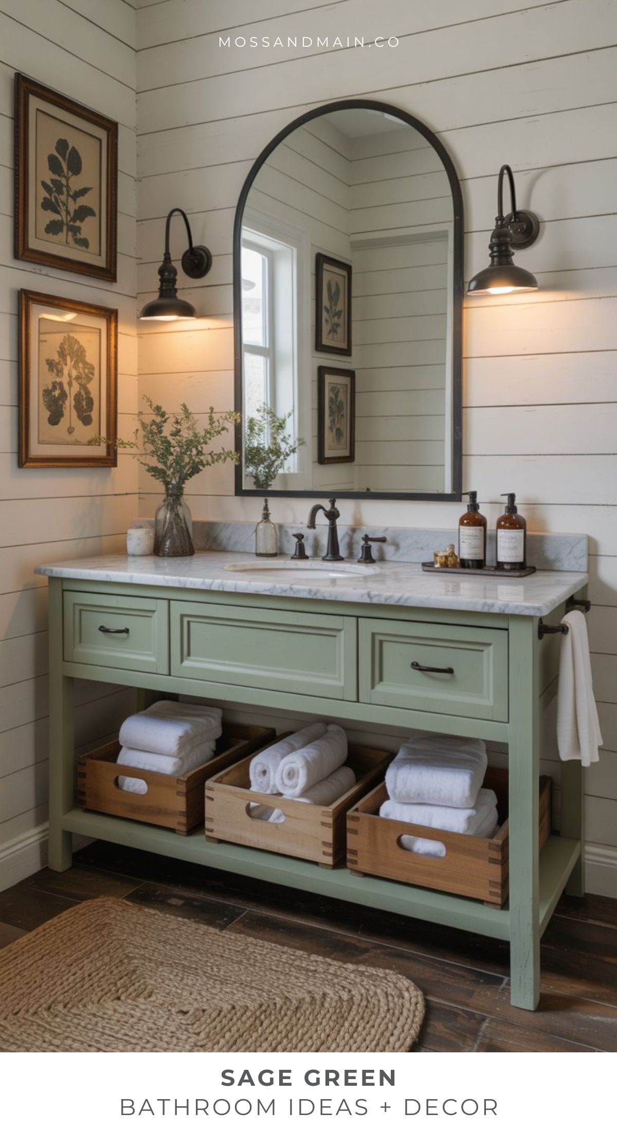 A sage green bathroom vanity with a marble top features two drawers and open shelves with wooden baskets of rolled towels. Above, an arch mirror, wall sconces, and framed botanical prints accent white shiplap walls—perfect bathroom ideas.