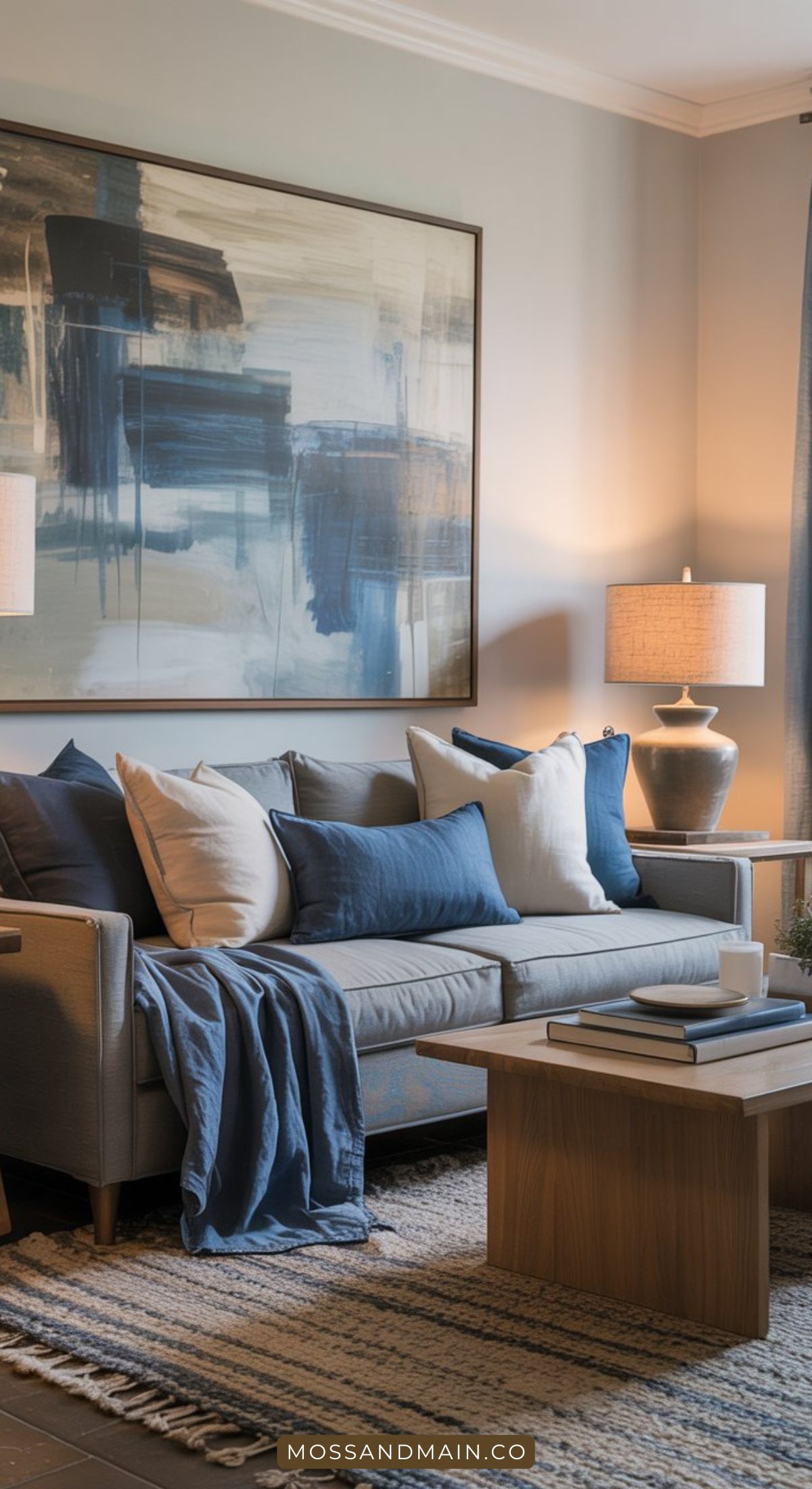 A cozy gray couch living room with blue and white pillows, a blue throw, a wooden coffee table, two table lamps, and a large abstract painting. Neutrals and textured rug complete this inviting living room decor.