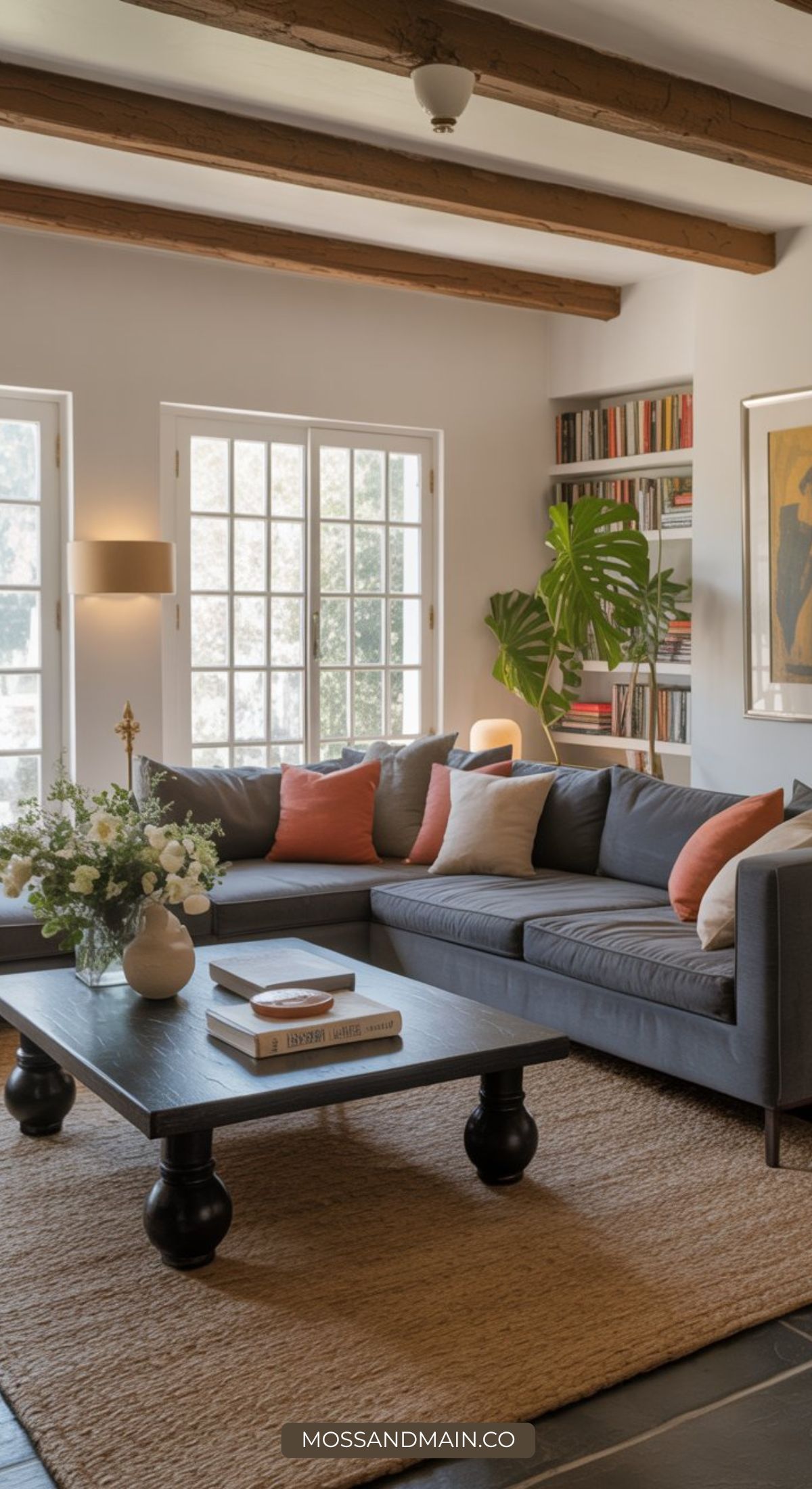 A cozy grey sofa living room with coral and cream pillows, a wooden coffee table, bookshelves, potted plants, large windows, and exposed ceiling beams. Warm light fills the inviting, stylish space.