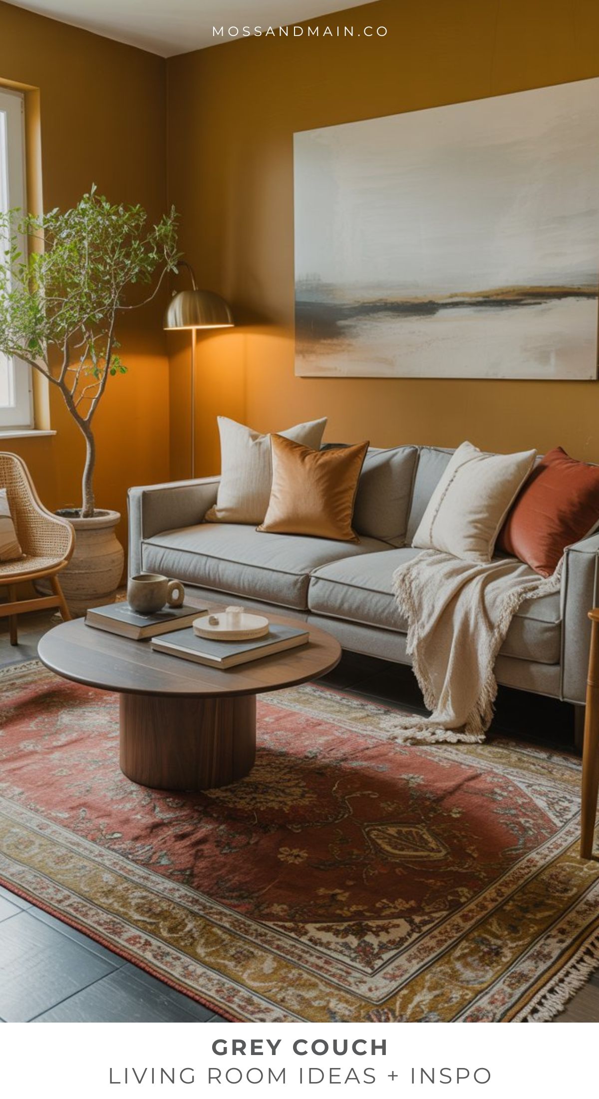 A cozy grey sofa living room features earth-toned pillows, a round wooden coffee table, a patterned red rug, large wall art, a tall gold floor lamp, and a wicker chair near a window with a potted plant.