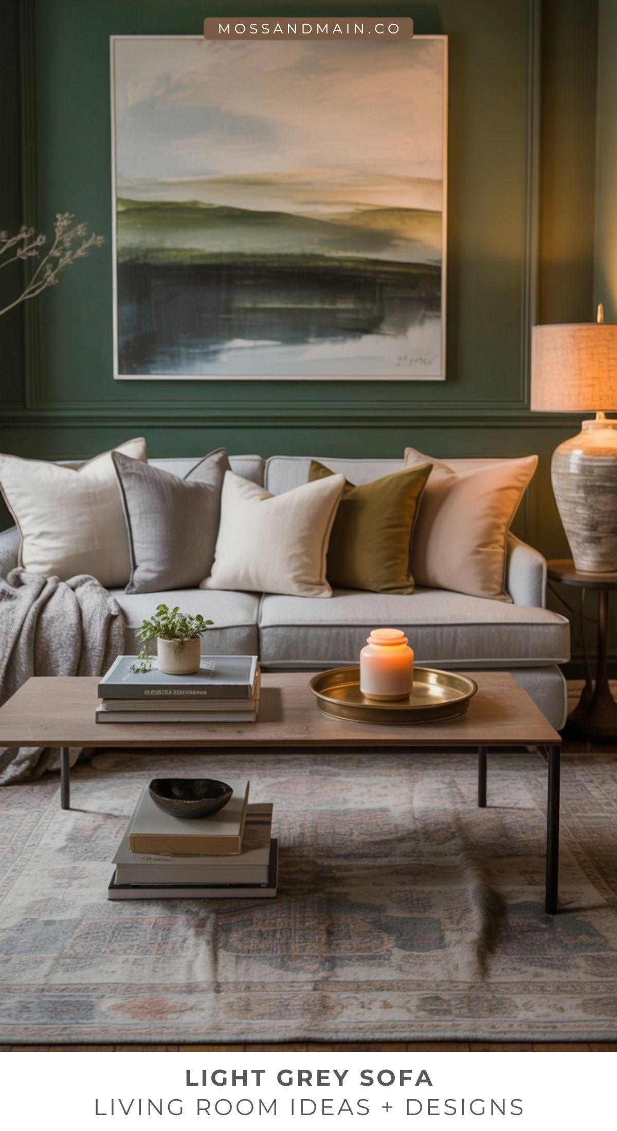 A cozy living room featuring a light gray couch, assorted neutral and tan pillows, a wooden coffee table with books, a plant, candle, and brass tray, set against a green wall with a large abstract painting above—perfect for gathering living room ideas.