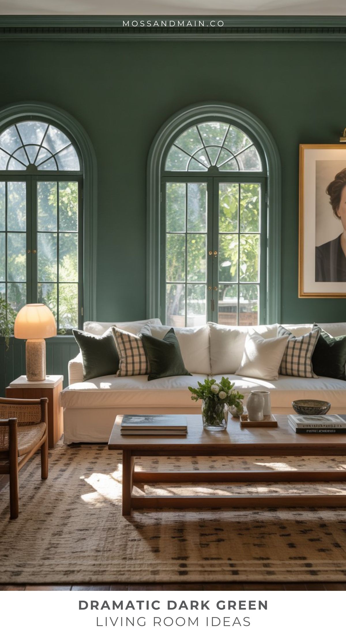 Elegant living room with dark forest green walls, tall arched windows, a white sofa with assorted pillows, a wooden coffee table with books, a lamp, and a large portrait. Sunlight filters in, creating the perfect moody room straight from a designer catalog.