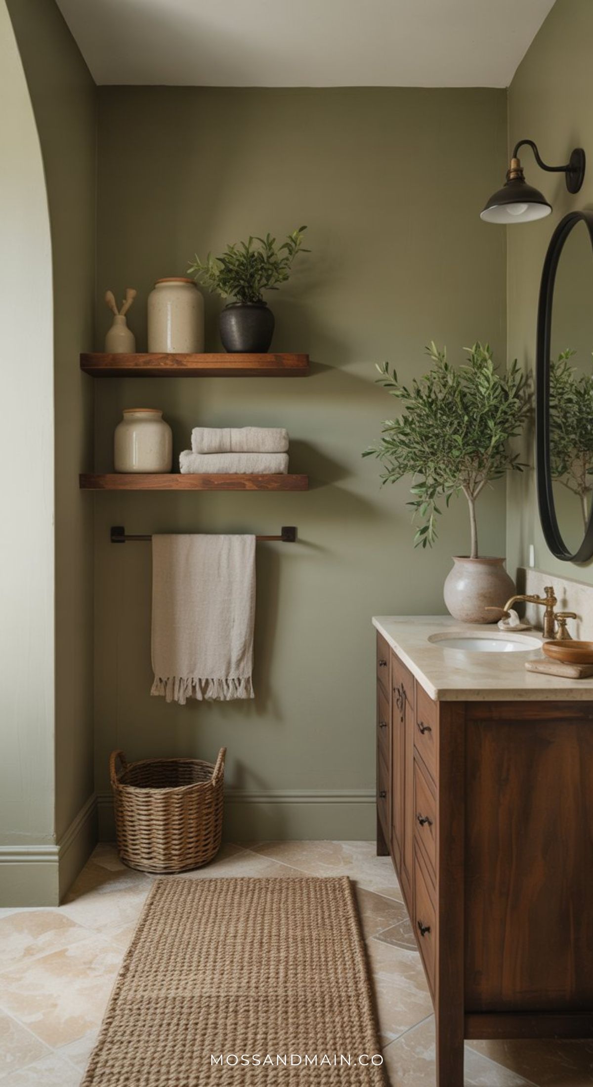 A cozy bathroom with sage green walls, wooden shelves holding jars and decor, a round mirror, a sink with a wooden vanity, a potted plant, a towel, a basket, and a woven rug on tile floor—perfect for timeless bathroom inspiration.