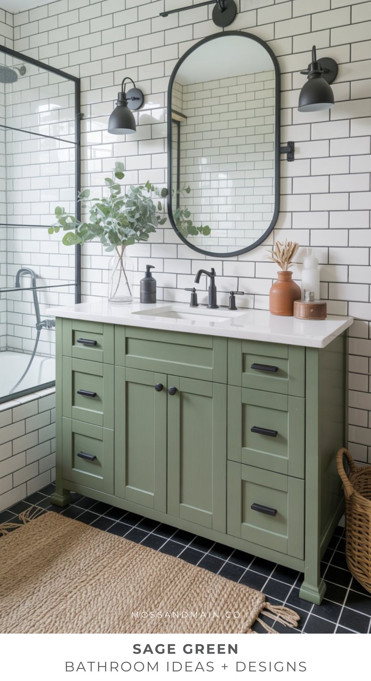 A cozy sage green bathroom vanity with black hardware sits against white subway tile walls, below an oval mirror and black wall sconces. For more bathroom ideas, decor includes plants, neutral bottles, and a woven rug on a black tile floor.