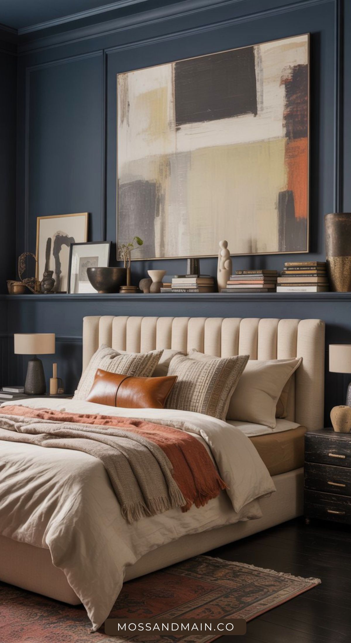 A modern bedroom with a beige upholstered bed, rust and neutral bedding, and a large abstract painting on a dark blue wall. Decorative objects and books are arranged on a shelf above the bed.