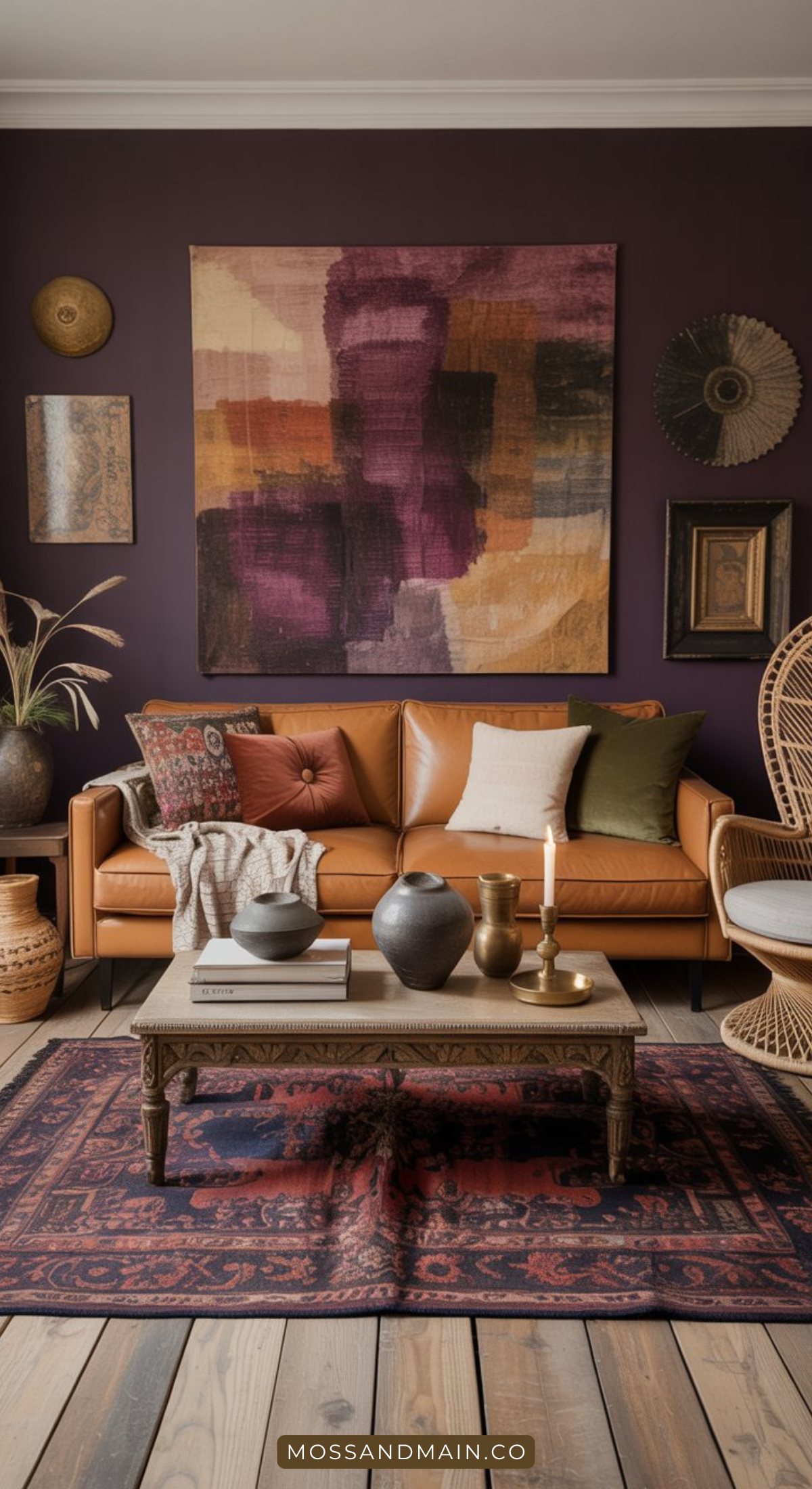 A cozy living room with a tan leather sofa, colorful pillows, and a woven chair. Embracing moody living room style, deep purple walls display eclectic art above a wooden coffee table with vases, books, and candles on a patterned rug.