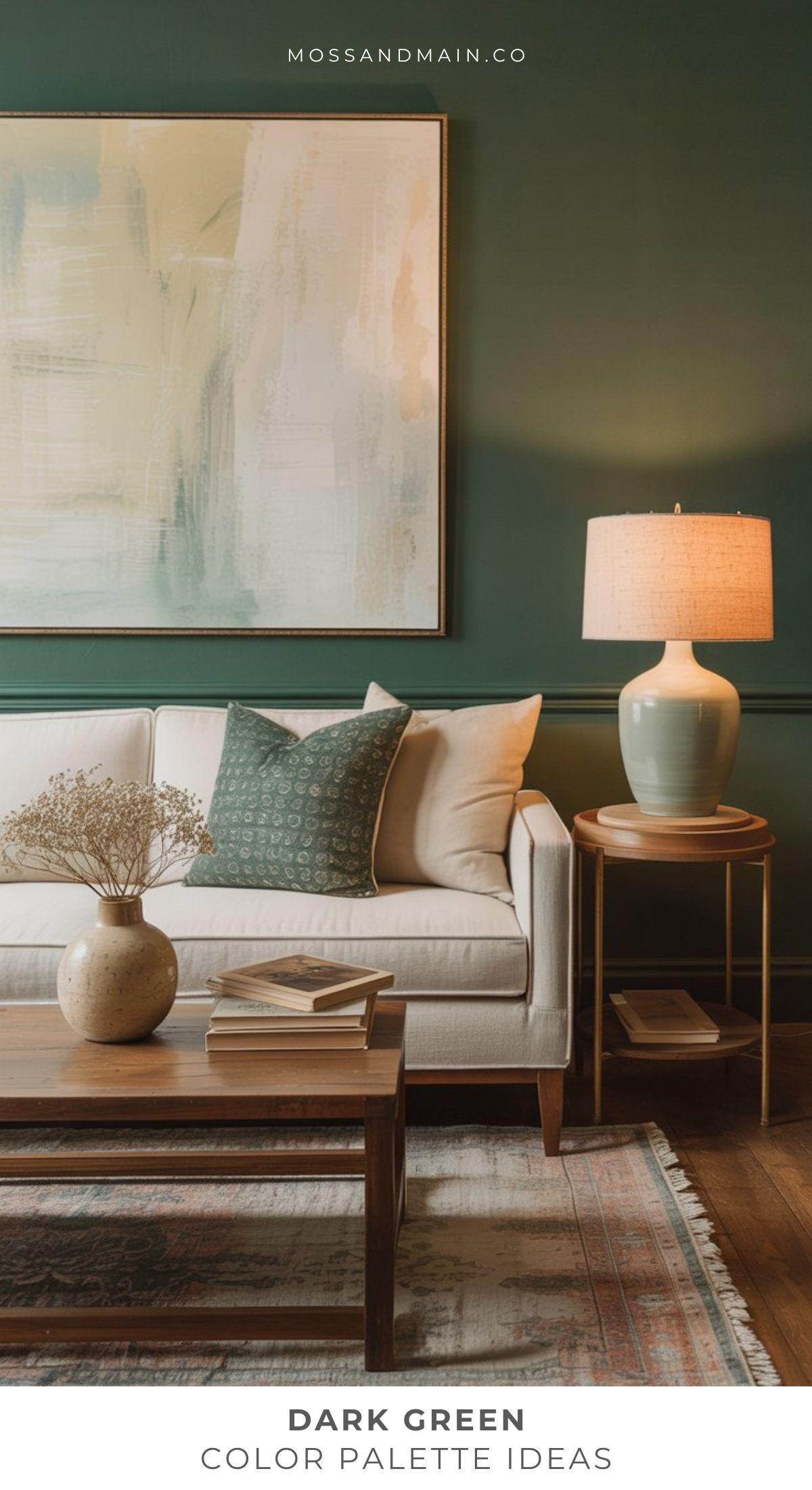 A living room with a dark green color scheme features matching walls, a white sofa with green and patterned pillows, a wooden coffee table topped with books and a vase, a side table with a lamp, and a large abstract painting above the sofa.