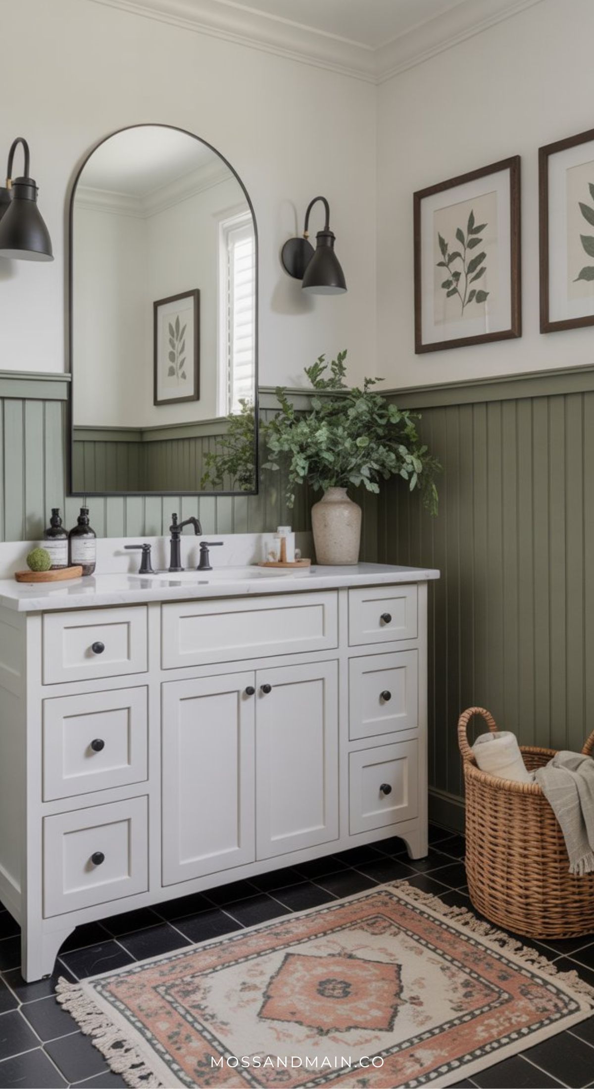 A modern, timeless bathroom with a white vanity, black hardware, and a large arched mirror. Sage green wainscoting lines the walls decorated with botanical prints. Towels in a basket and a patterned rug sit on the black tile floor.