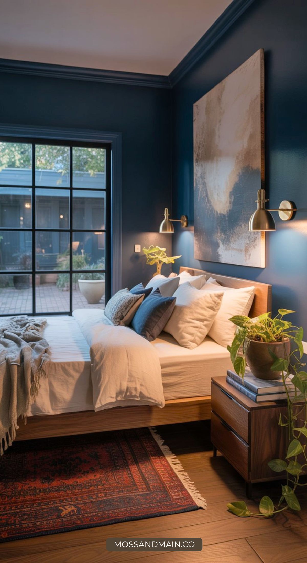 A stylish blue boho bedroom with dark blue walls, a large abstract painting above the bed, white bedding, modern wall sconces, and a wooden nightstand with books and a plant. Large windows overlook a patio with greenery.