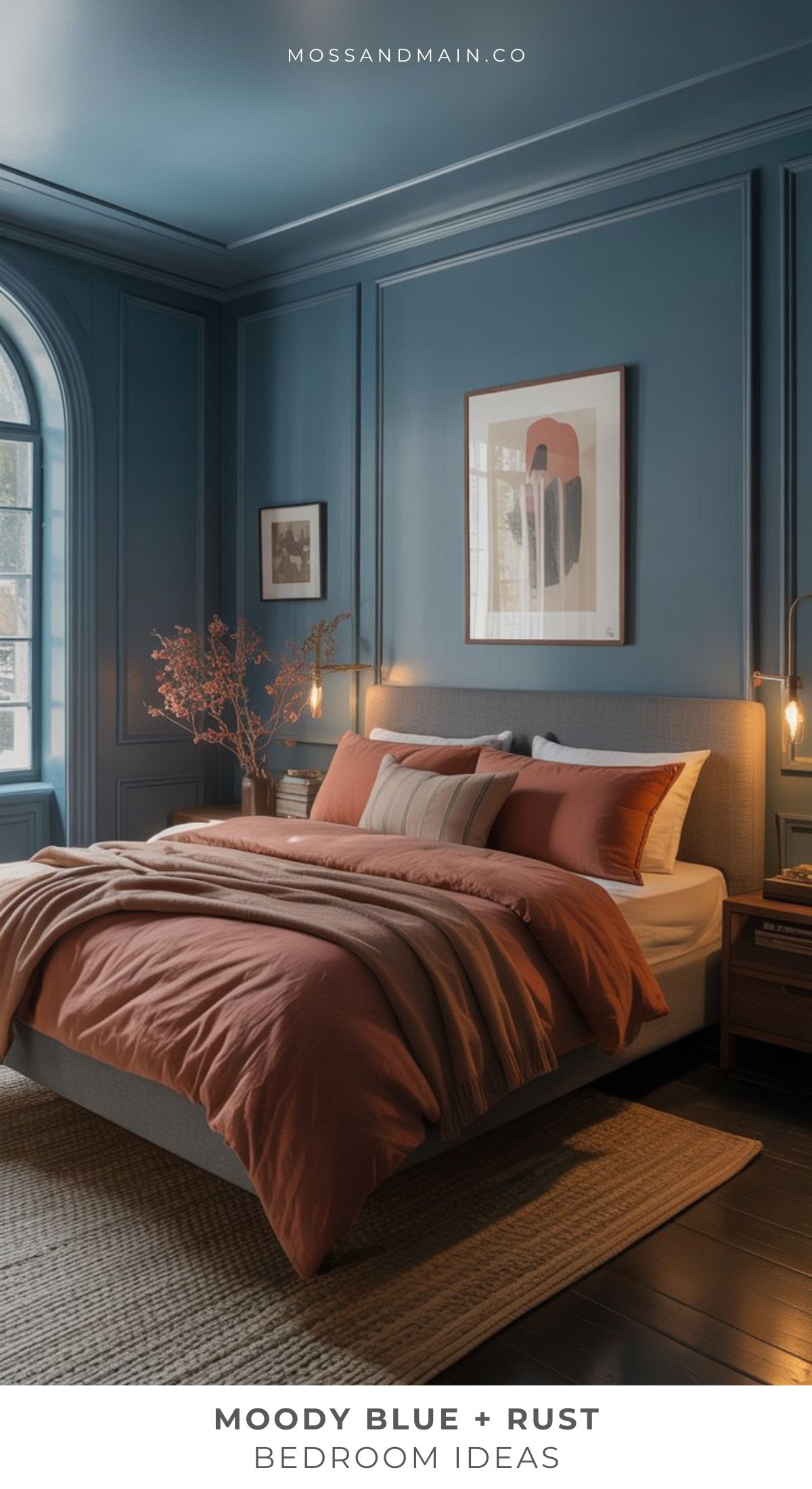 A stylish dusty blue bedroom with moody walls and ceiling features a gray bed with rust and beige bedding. Framed abstract art hangs above, while natural light from the window highlights a woven rug—perfect for earthy bedroom ideas.