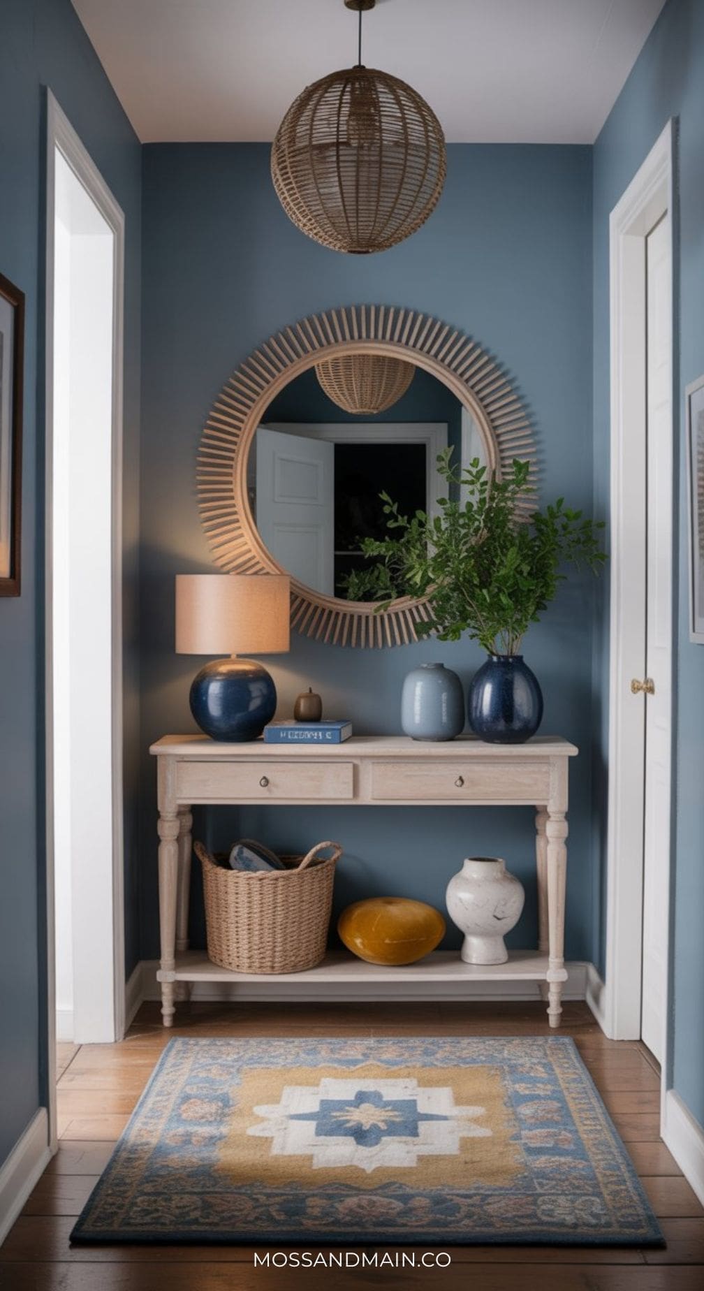 A cozy hallway with blue walls and a touch of mustard color palette, a round mirror, wooden console table with a lamp, vases, books, a basket, and green branches. A patterned rug lies on the wooden floor. Warm and inviting decor.
