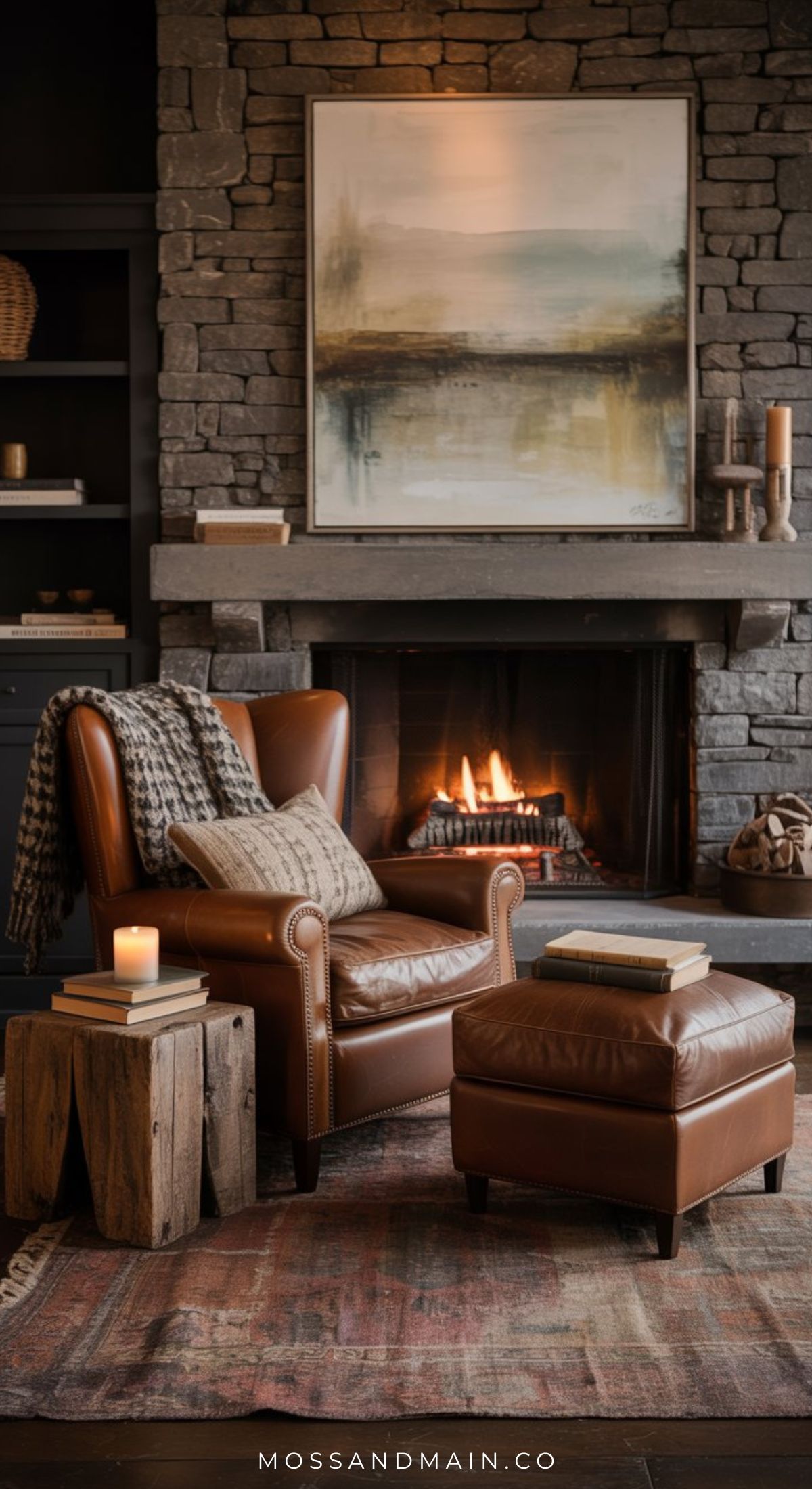 A cozy reading nook with a leather armchair, ottoman, knit blanket, wooden side tables, and a lit candle sits in front of a stone fireplace with a modern painting above it—your little escape in warm, inviting tones.