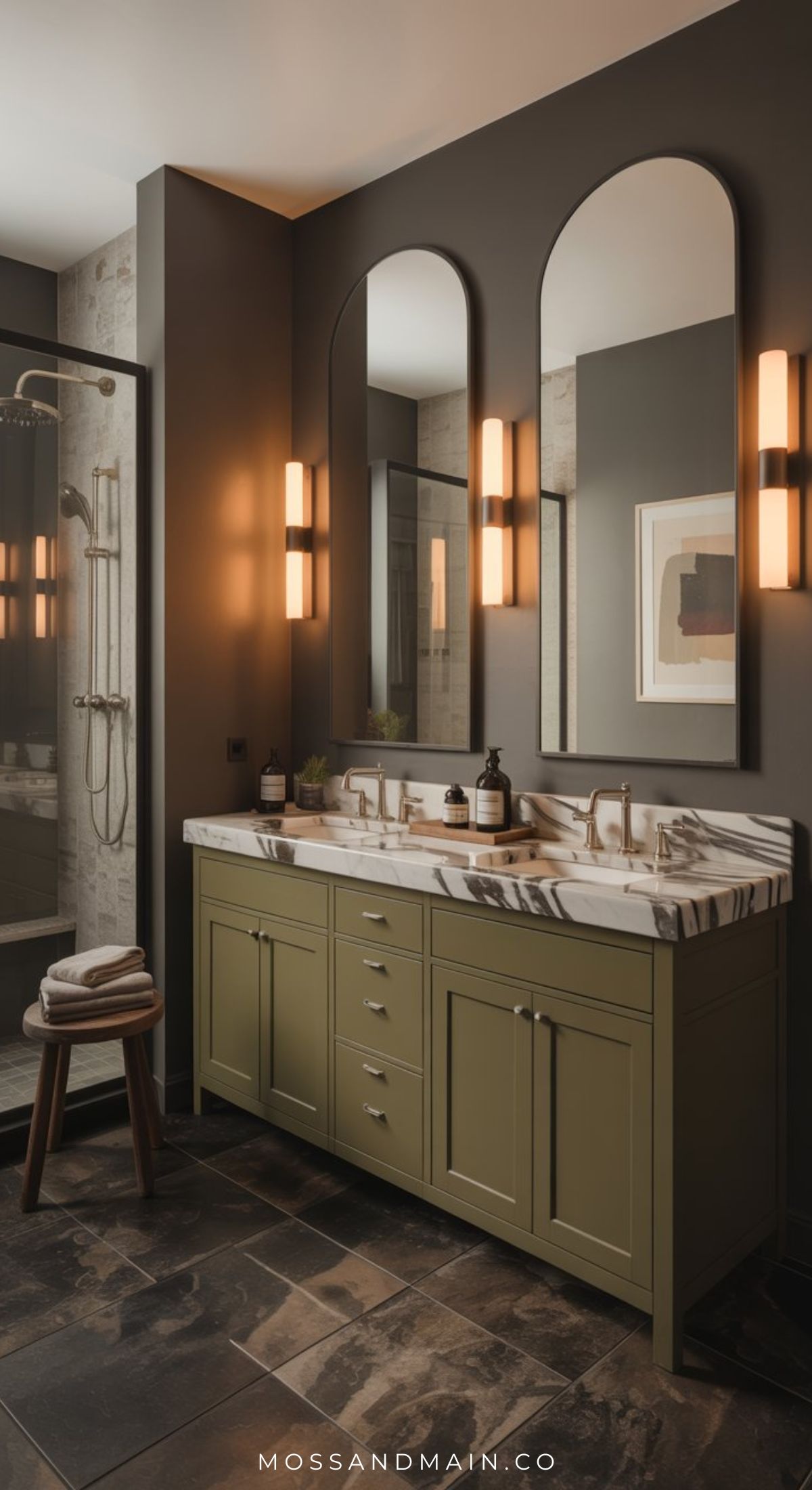 A modern bathroom with an olive green double vanity, marble countertop, two tall mirrors, warm wall sconces, and a glass-enclosed shower; towels are stacked on a wooden stool nearby.