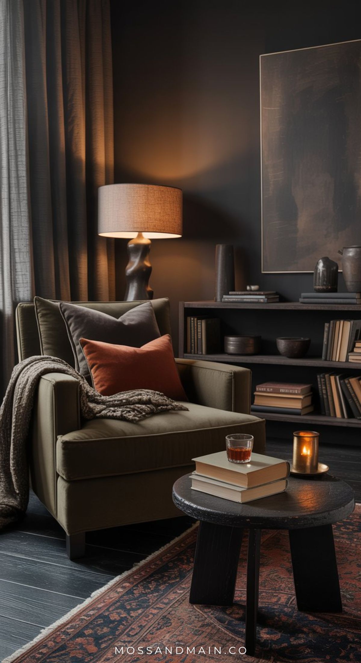 A cozy living room with a dark armchair, orange and brown cushions, a throw blanket, a floor lamp, shelves filled with books and ceramics, and a small round table with books, a candle, and a glass of drink.