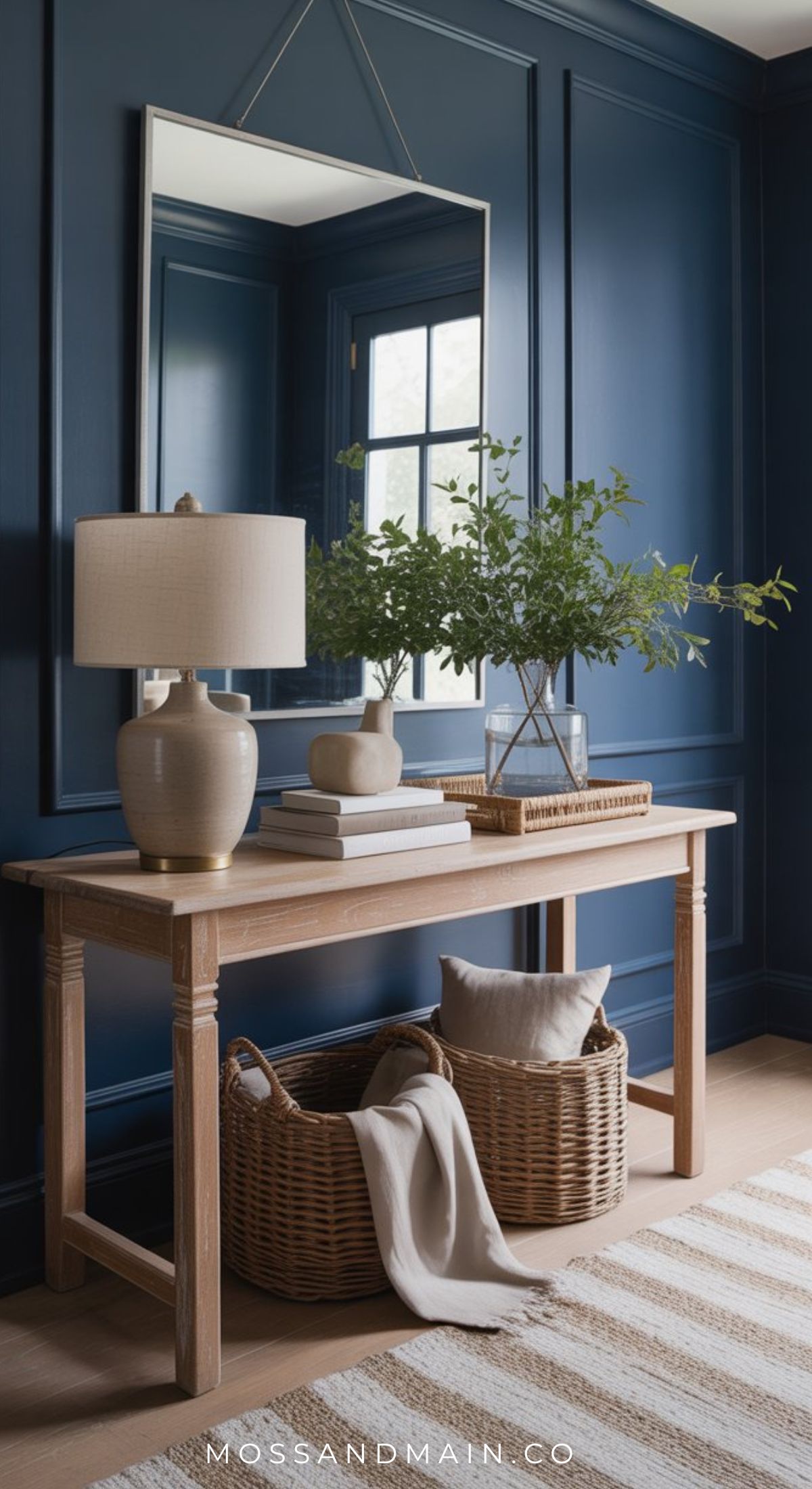 A wooden console table sits against a dark blue paneled wall, topped with a beige lamp, books, and a glass vase with green branches. Two woven baskets and a striped blanket rest below—perfect inspiration for warm entryway style. A large mirror hangs above.