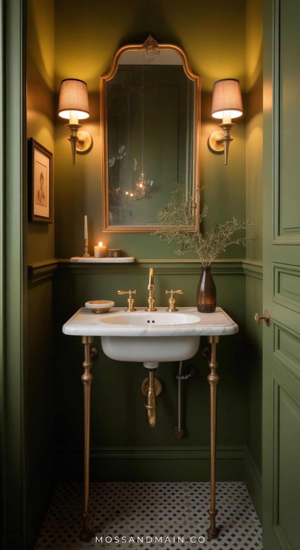 A stylish olive green bathroom with gold accents features a white sink, brass fixtures, a gold-framed mirror, wall sconces, a vase with branches, a candle, and black-and-white hexagon floor tiles—perfect for moody bathroom ideas.