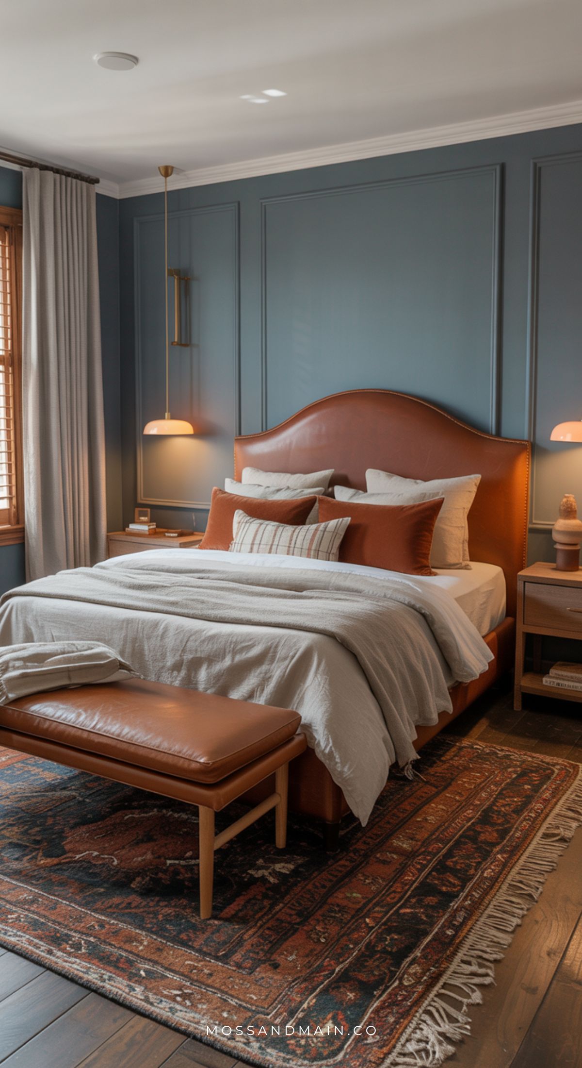 A cozy earthy vibe fills this bedroom with a brown leather bed frame, beige bedding, brown and white pillows, blue paneled walls, warm lighting, wooden nightstands, and a patterned rug—perfect for dusty blue bedroom inspiration.